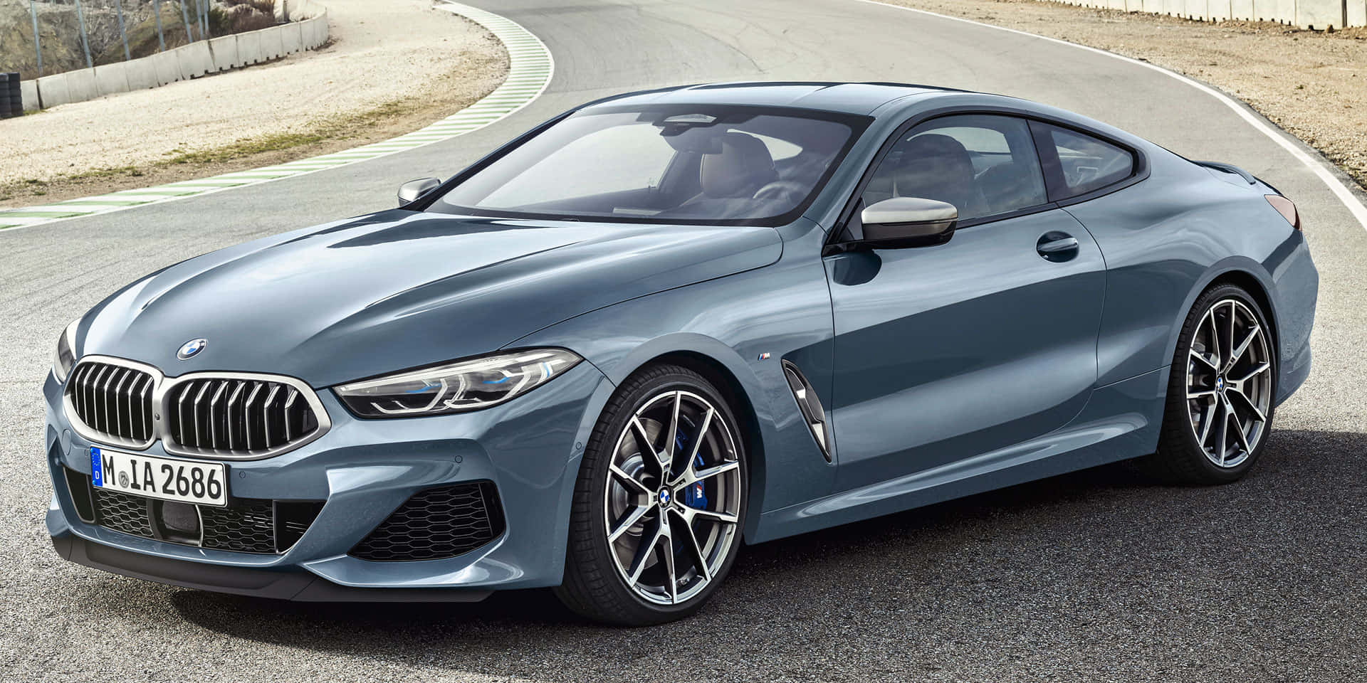 Bmw 8 Series 桌布 壁紙 — 一輛時尚的金属藍色BMW 8系轎跑車在明亮的日光下，沿著一條由礫石和綠樹環繞的彎曲瀝青道路滑行。