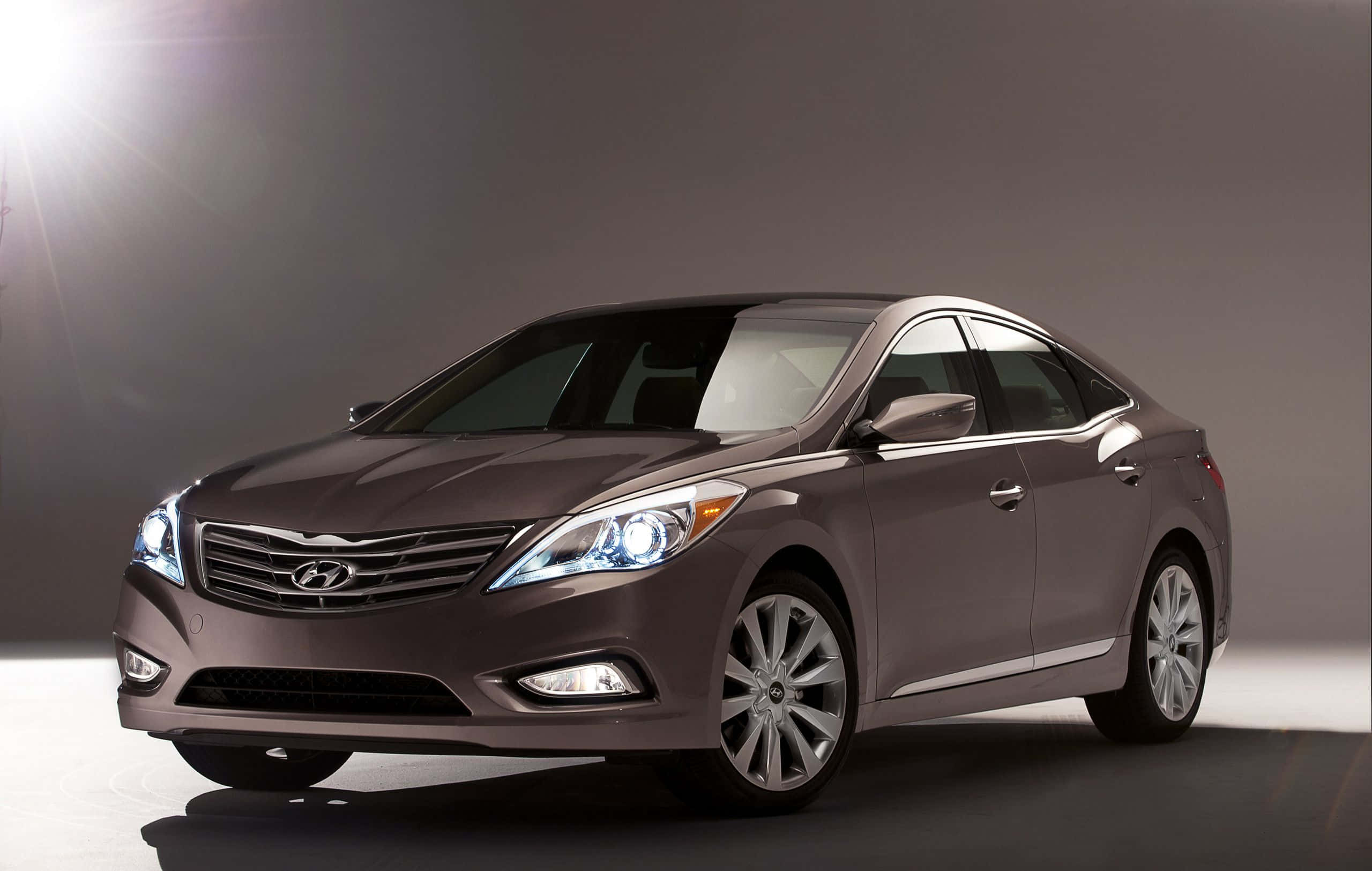 Hyundai Azera 桌布 壁紙 — 一輛時尚的金屬棕色轎車，配備發光的頭燈，在具有戲劇性燈光和漸變灰色背景的攝影棚中展示。