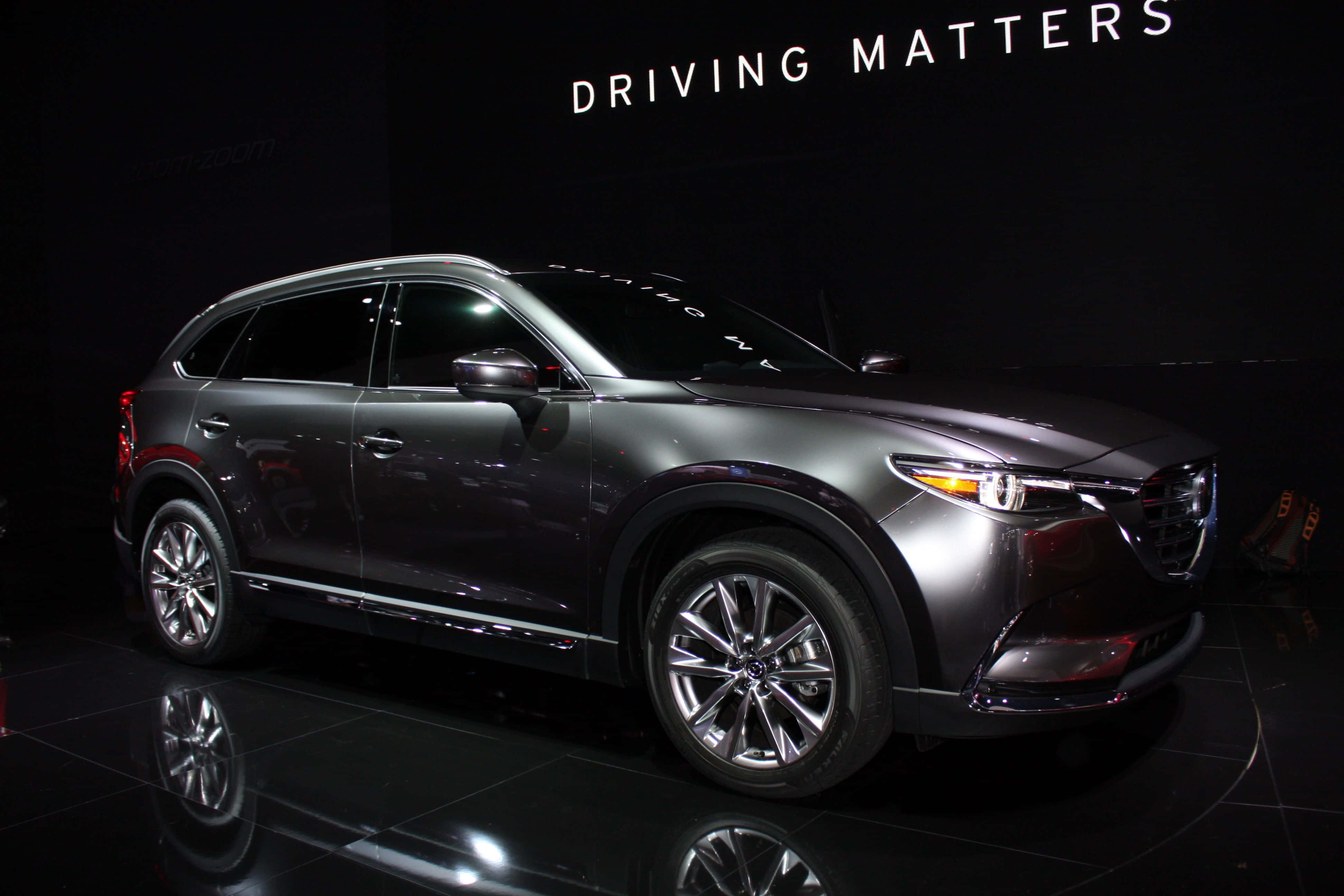 Mazda Cx-9 桌布 壁紙 — 一輛時尚的金屬灰色萬事得SUV在聚光燈下於反射黑色舞台上閃耀，上方有'DRIVING MATTERS'標牌。