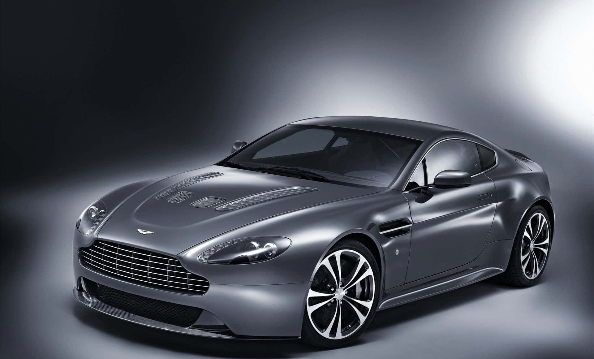 Aston Martin V12 Vantage 桌布 壁紙 — 一輛時尚的銀色 Aston Martin 跑車在戲劇性的攝影棚燈光下，於深色漸變背景前閃耀光芒。