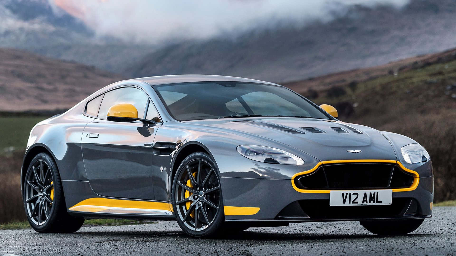Aston Martin V12 Vantage 桌布 壁紙 — 一輛時尚的銀色 Aston Martin V12 AMR 帶有黃色點綴，停泊在碎石路上，背景是霧氣繚繞的山丘。