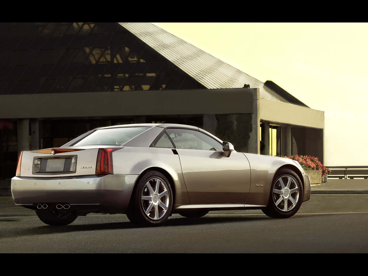 Cadillac Xlr 桌布 壁紙 — 一輛時尚的銀色 Cadillac XLR 雙門跑車停在瀝青路面上，旁邊是一座帶有斜屋頂和花槽的現代建築。