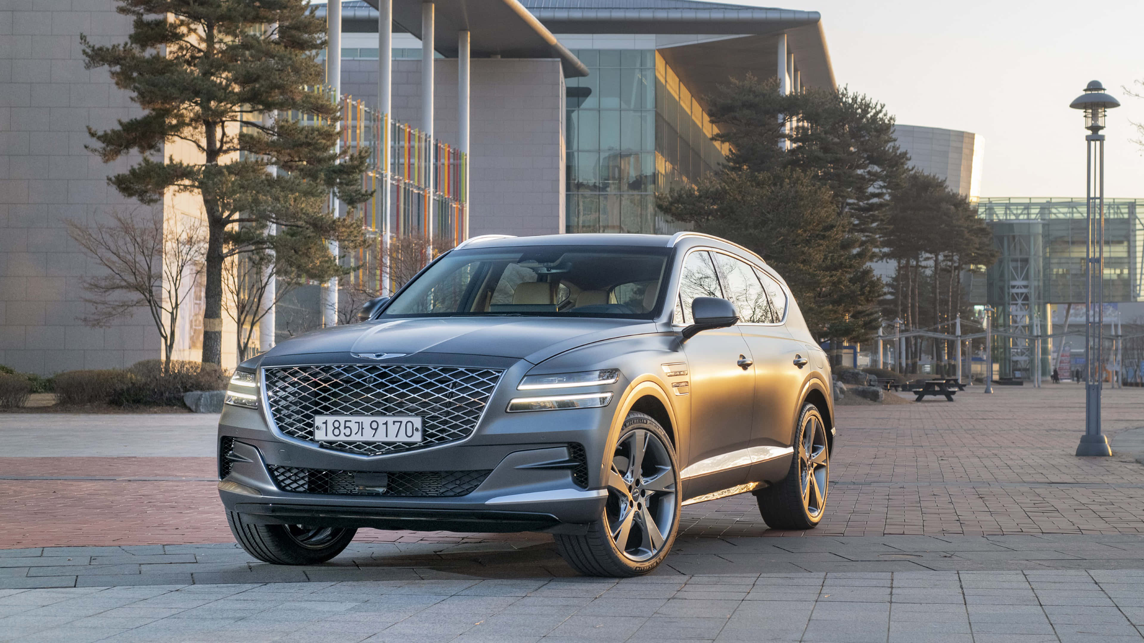 Genesis Gv80 桌布 壁紙 — 一輛時尚的銀色 Genesis GV80 SUV 停泊在鋪砌的廣場上，背景是現代建築和樹木，正值黃金時段。