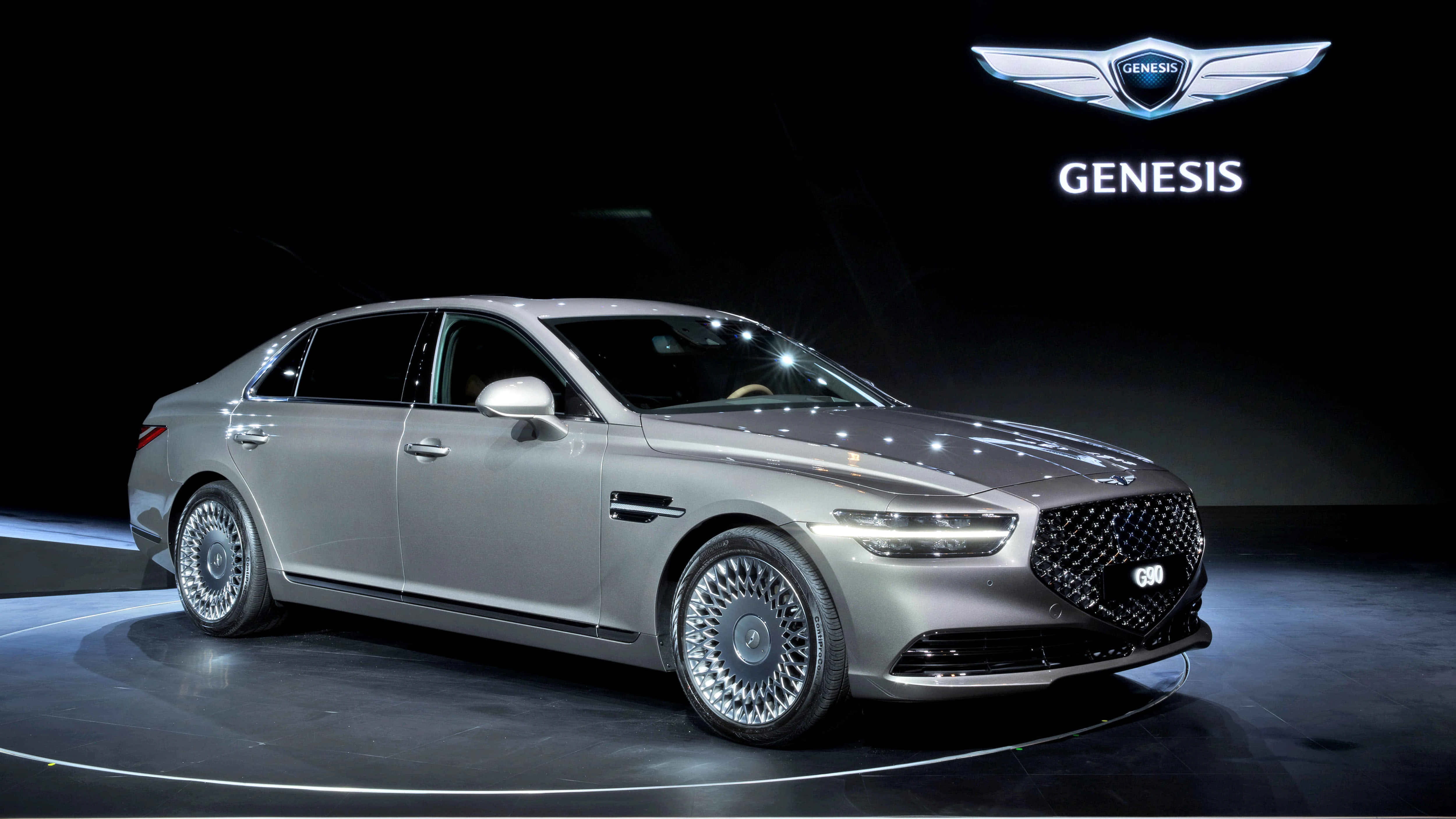 Genesis G90 桌布 壁紙 — 一輛時尚的銀色 Genesis 豪華轎車在攝影棚燈光下於黑暗舞台上閃耀，品牌標誌在上方發光。