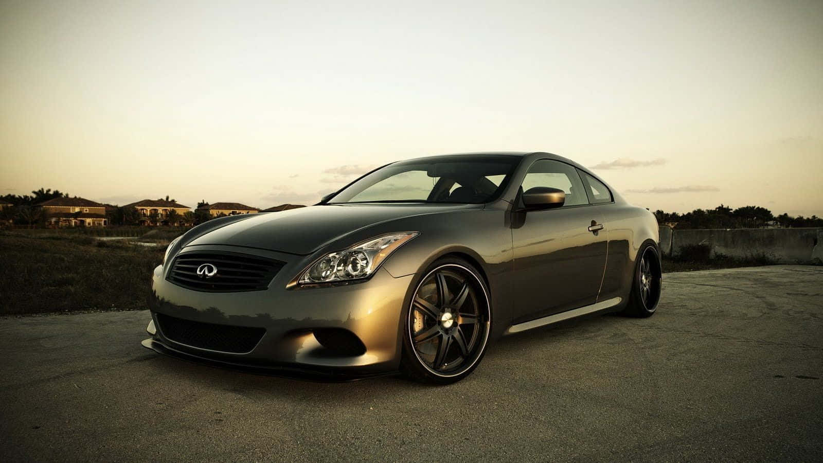 Infiniti G35 桌布 壁紙 — 一輛時尚的銀色 Infiniti 雙門轎跑車在金色夕陽下閃閃發光，停在空曠的瀝青停車場上，遠處有房屋。