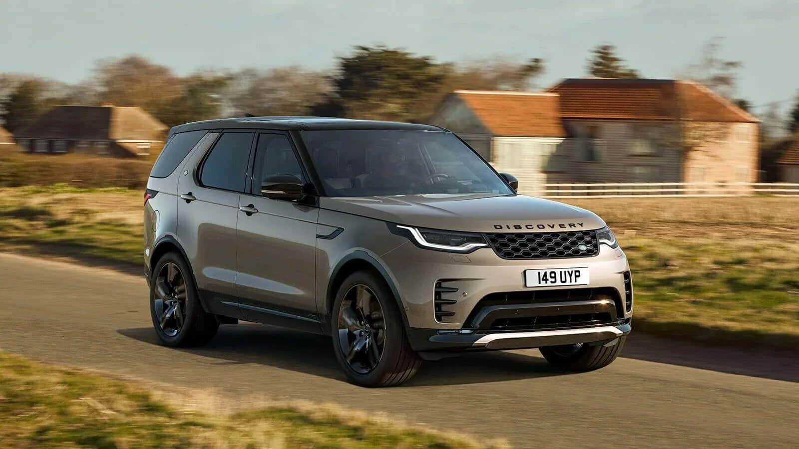 Land Rover Discovery 桌布 壁紙 — 一輛時尚的銀色 Land Rover Discovery 沿著鄉村道路行駛，經過質樸的石屋，天空晴朗。