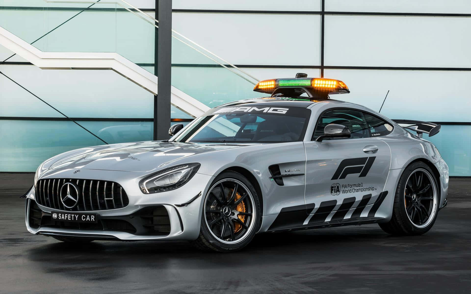 安全車在賽道上高速領跑 壁紙 — 一輛時尚的銀色 Mercedes-AMG GT 安全車，帶有閃爍的燈光，停在一棟現代玻璃建築前。