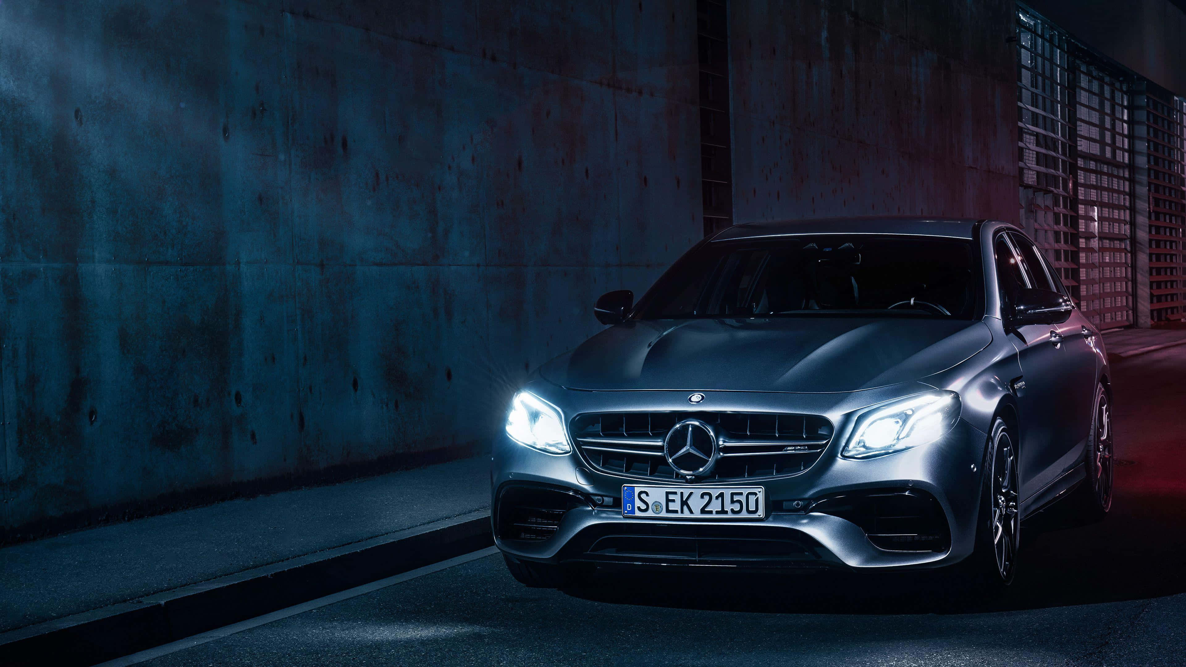 AMG E-63 S Mercedes Benz 4K 壁紙 — 一輛時尚的銀色 Mercedes-AMG E63 S 轎車，配備發光頭燈，停在光線昏暗的城市隧道中，帶有藍色和紅色的燈光點綴。