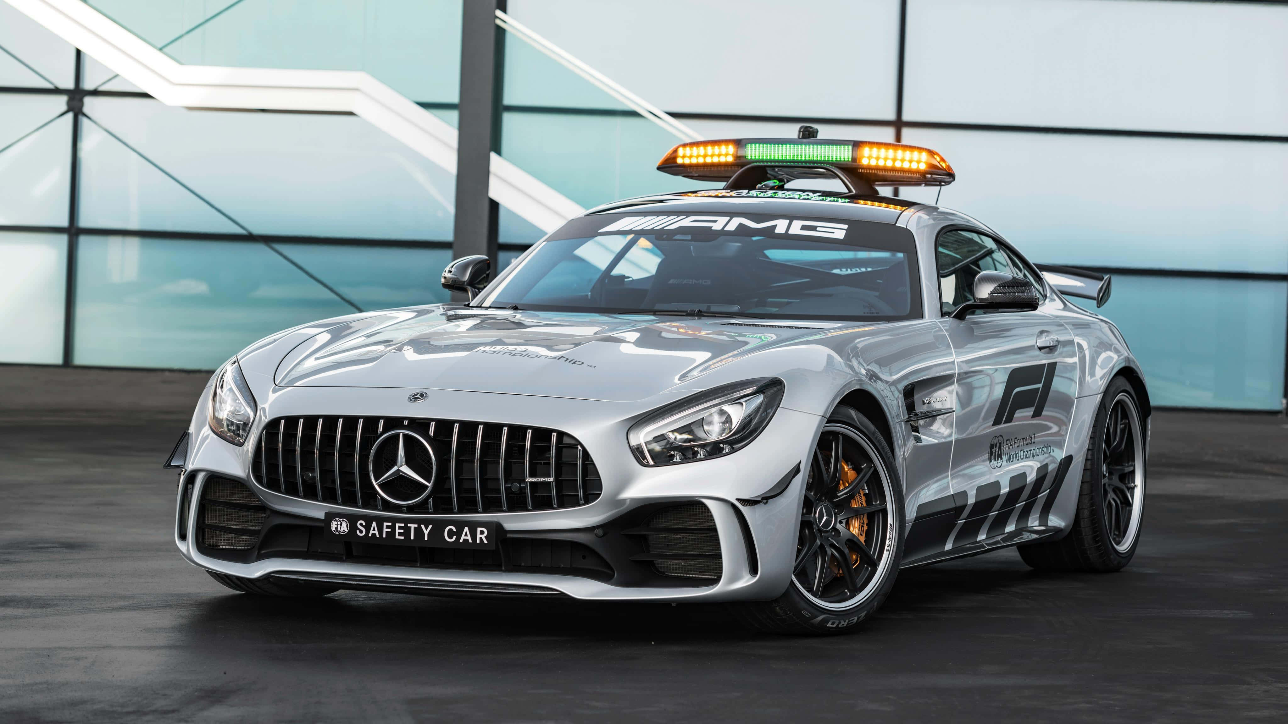 Safety Car 桌布 壁紙 — 一輛時尚的銀色 Mercedes-AMG GT 安全車，帶有閃爍的燈光，停在一棟現代玻璃建築前。