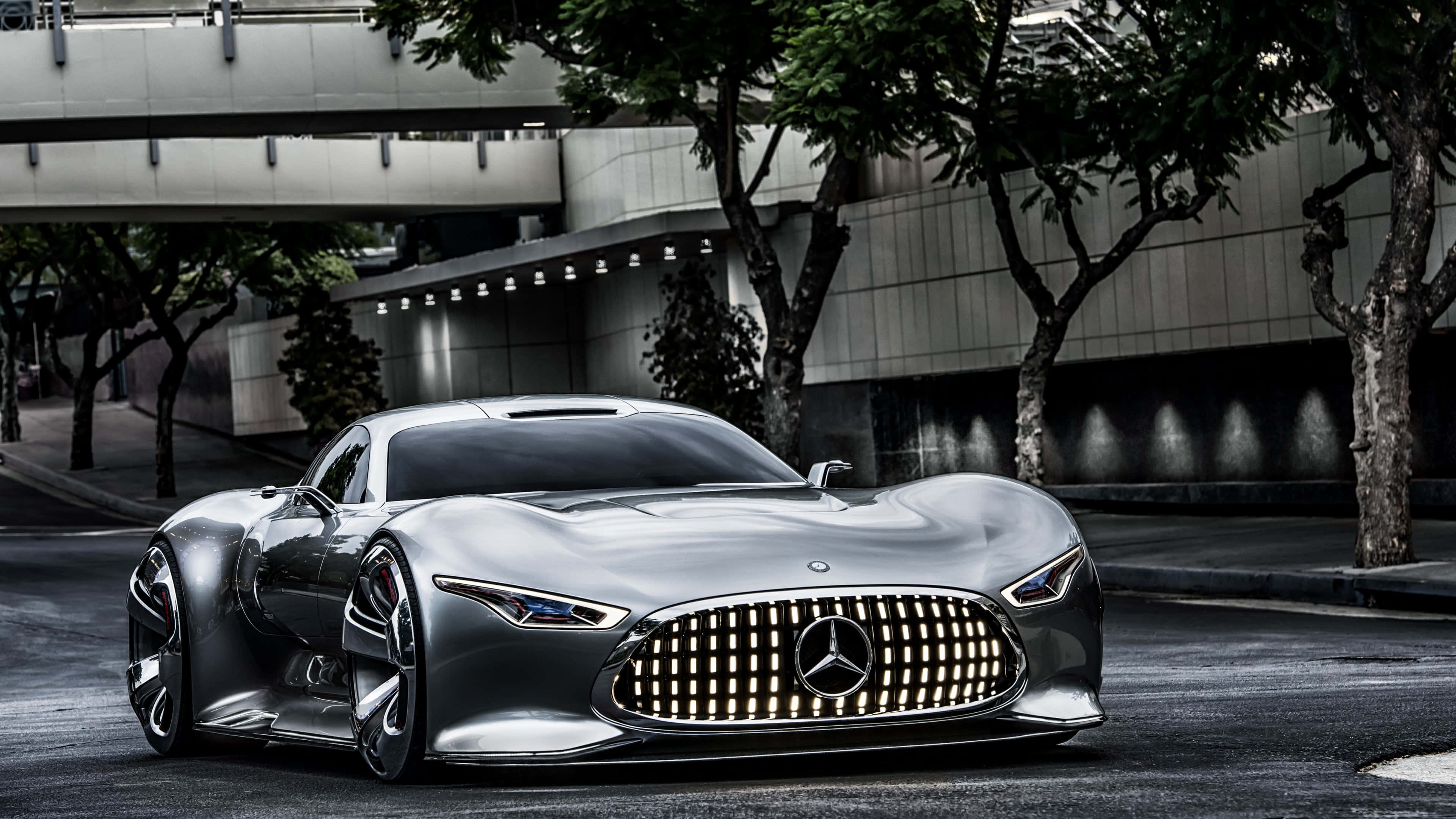 4K Mercedes 桌布 壁紙 — 一輛時尚的銀色 Mercedes-Benz 概念車在都市街燈下，在枝葉繁茂的樹木和混凝土建築旁閃閃發光。