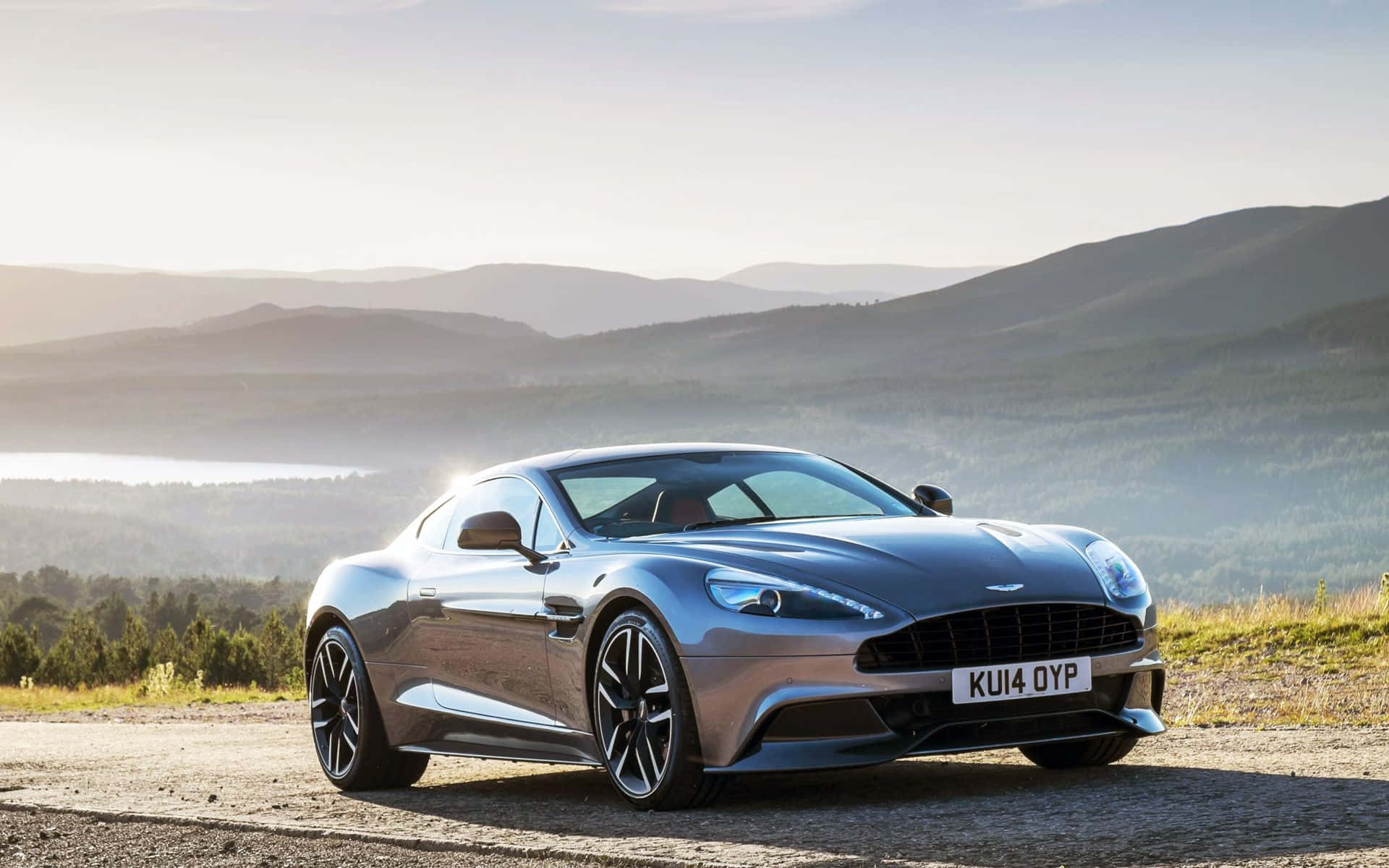 Aston Martin Db9 桌布 壁紙 — 一輛時尚的銀色Aston Martin跑車停在碎石路上，背景是連綿起伏的山丘和遠處的湖泊，天空柔和晨光。