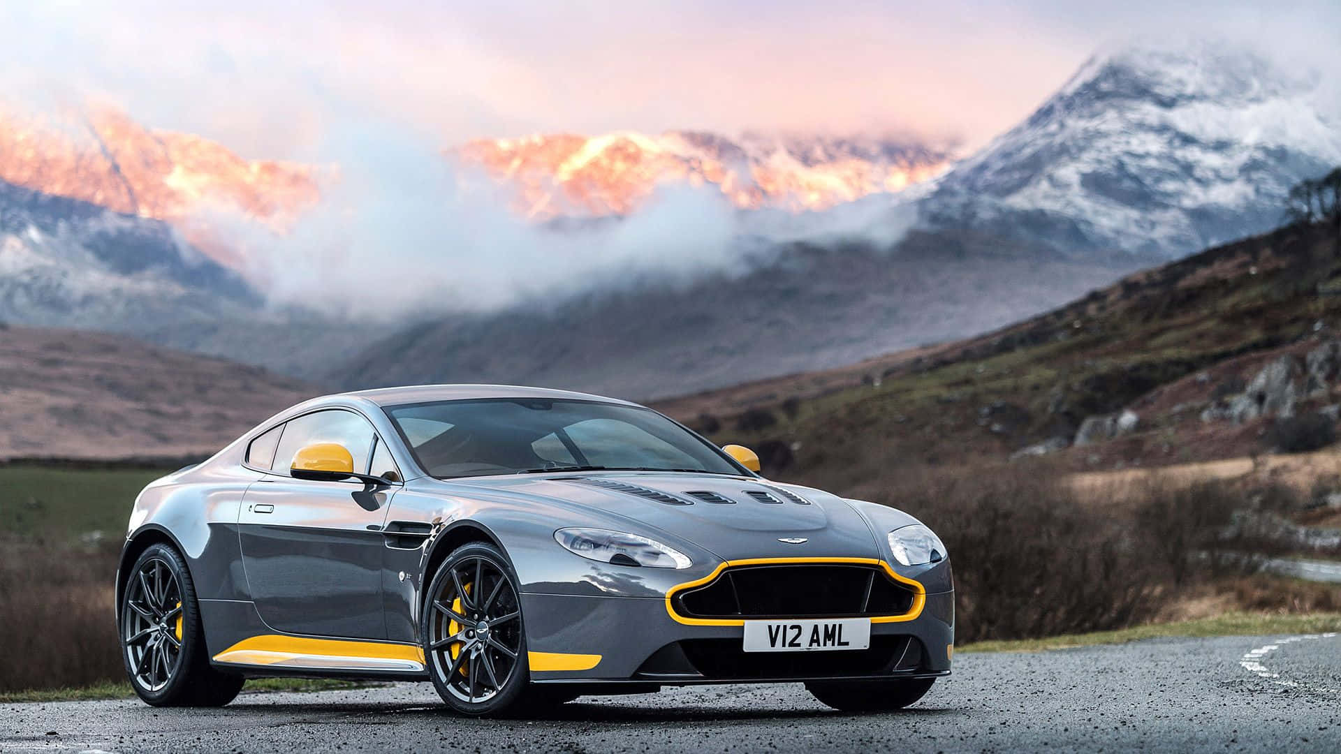 Aston Martin V12 Vantage 桌布 壁紙 — 一輛時尚的銀色Aston Martin帶有黃色點綴，停在碎石路上，背景是夕陽下閃耀的雪山。