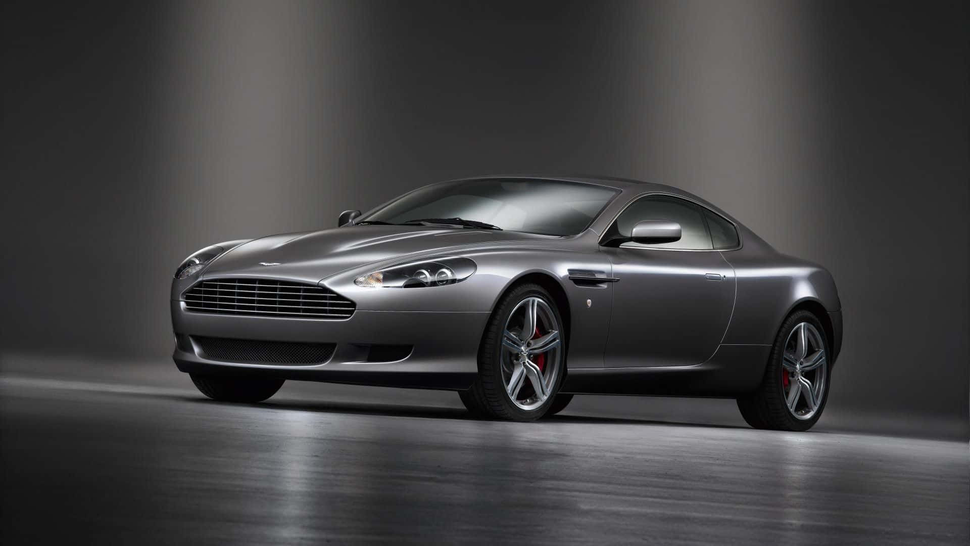 Aston Martin Db9 桌布 壁紙 — 一輛時尚的銀色Aston Martin跑車在戲劇性的工作室燈光下，在深色反光地板上滑行。
