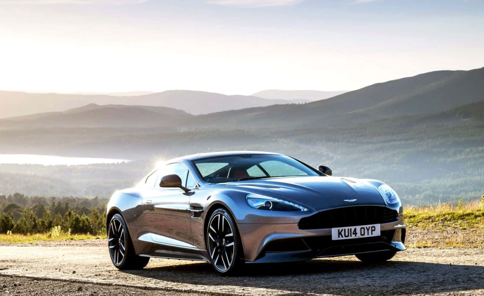 Aston Martin Vanquish 桌布 壁紙 — 一輛時尚的銀色Aston Martin跑車停在碎石路上，背景是日出時分連綿起伏的山丘和霧氣瀰漫的湖泊。