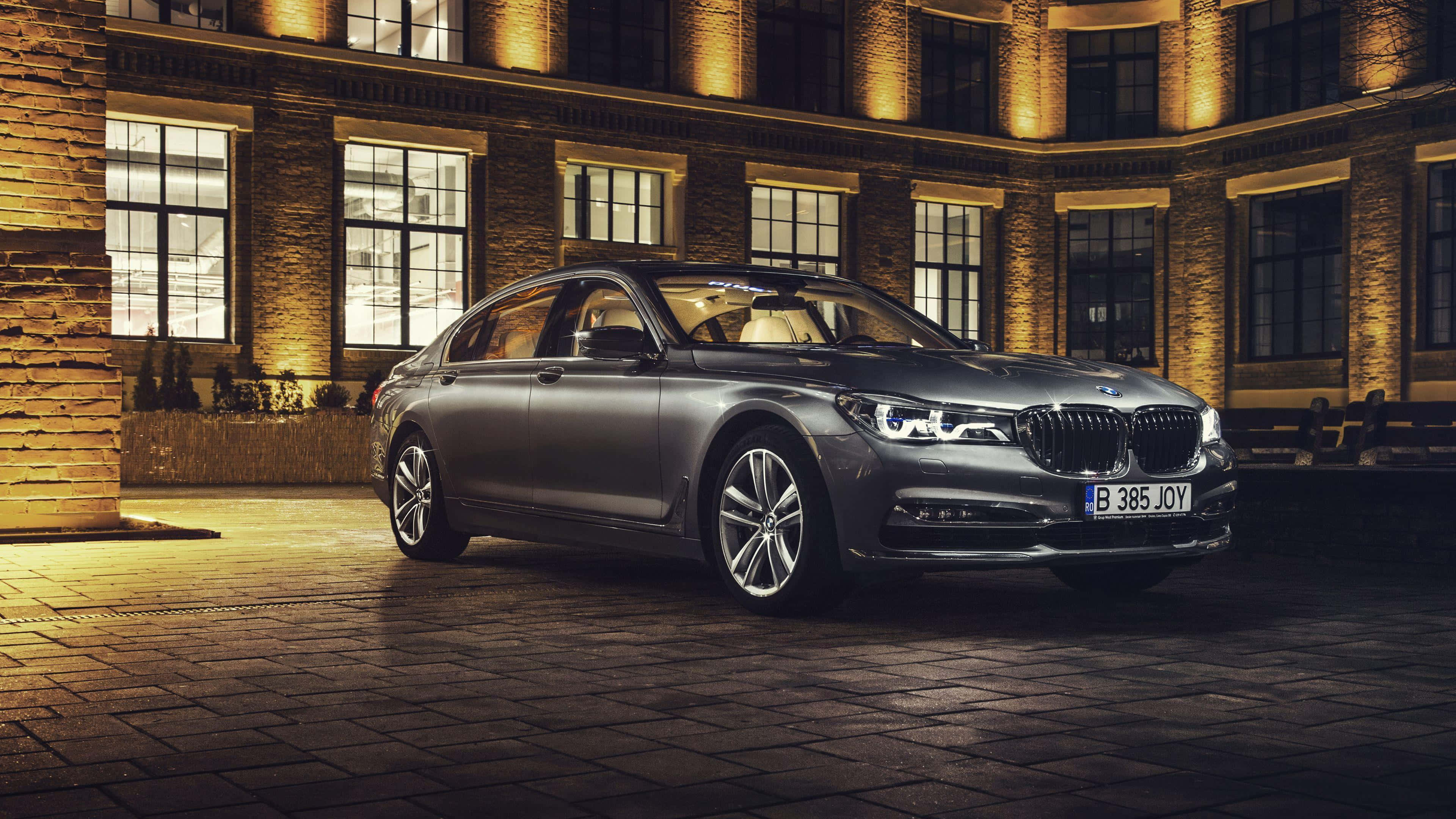 Bmw 7 Series 桌布 壁紙 — 一輛時尚的銀色BMW 7系轎車在夜間停泊在鵝卵石庭院，被身後歷史悠久的磚砌建築散發的溫暖金色燈光照亮。
