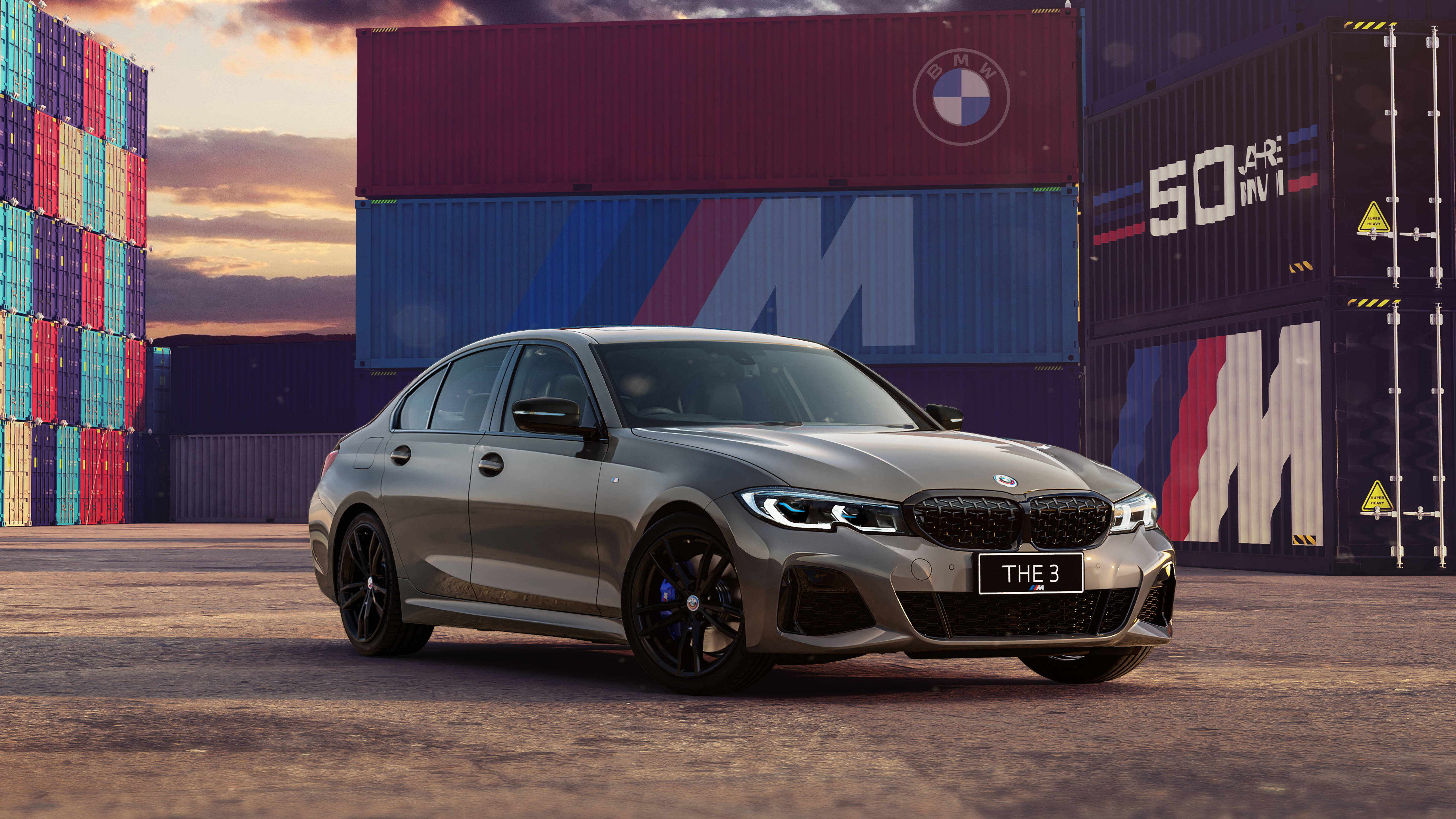Bmw 桌布 壁紙 — 一輛時尚的銀色BMW M3轎車在日落時停放在色彩繽紛的貨櫃之間。