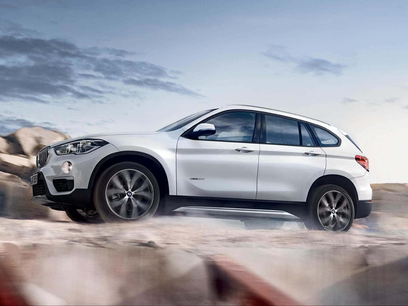 Bmw X1 桌布 壁紙 — 一輛時尚的銀色BMW X1 SUV在多雲的天空下，穿越塵土飛揚的沙漠景觀。