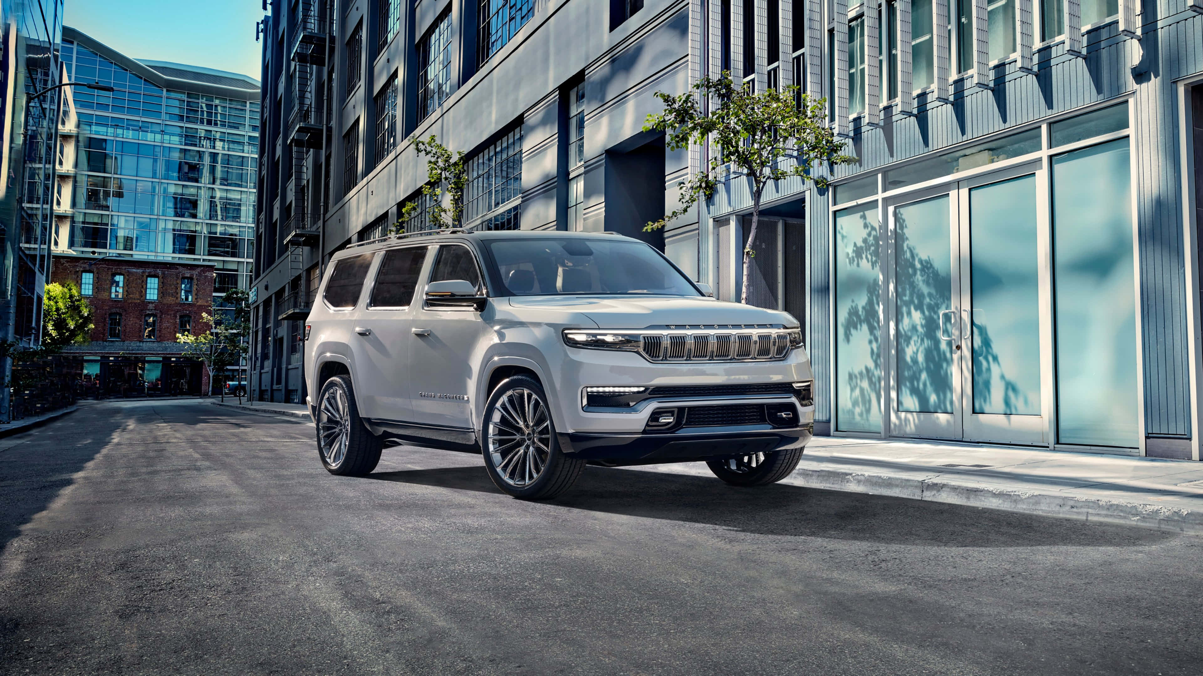 Jeep Wagoneer 桌布 壁紙 — 一輛時尚的銀色Jeep Grand Wagoneer停泊在現代城市街道上，兩旁是玻璃和鋼鐵建築，在明亮的日光下。