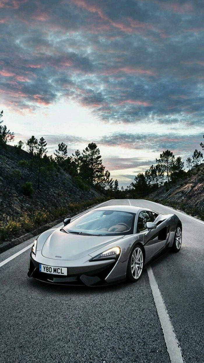 McLaren 570S Adobe Photoshop 壁紙 — 一輛時尚的銀色McLaren跑車沿著蜿蜒的山路行駛，在戲劇性的暮色天空下，天空被粉紅色和紫色的雲彩塗抹。