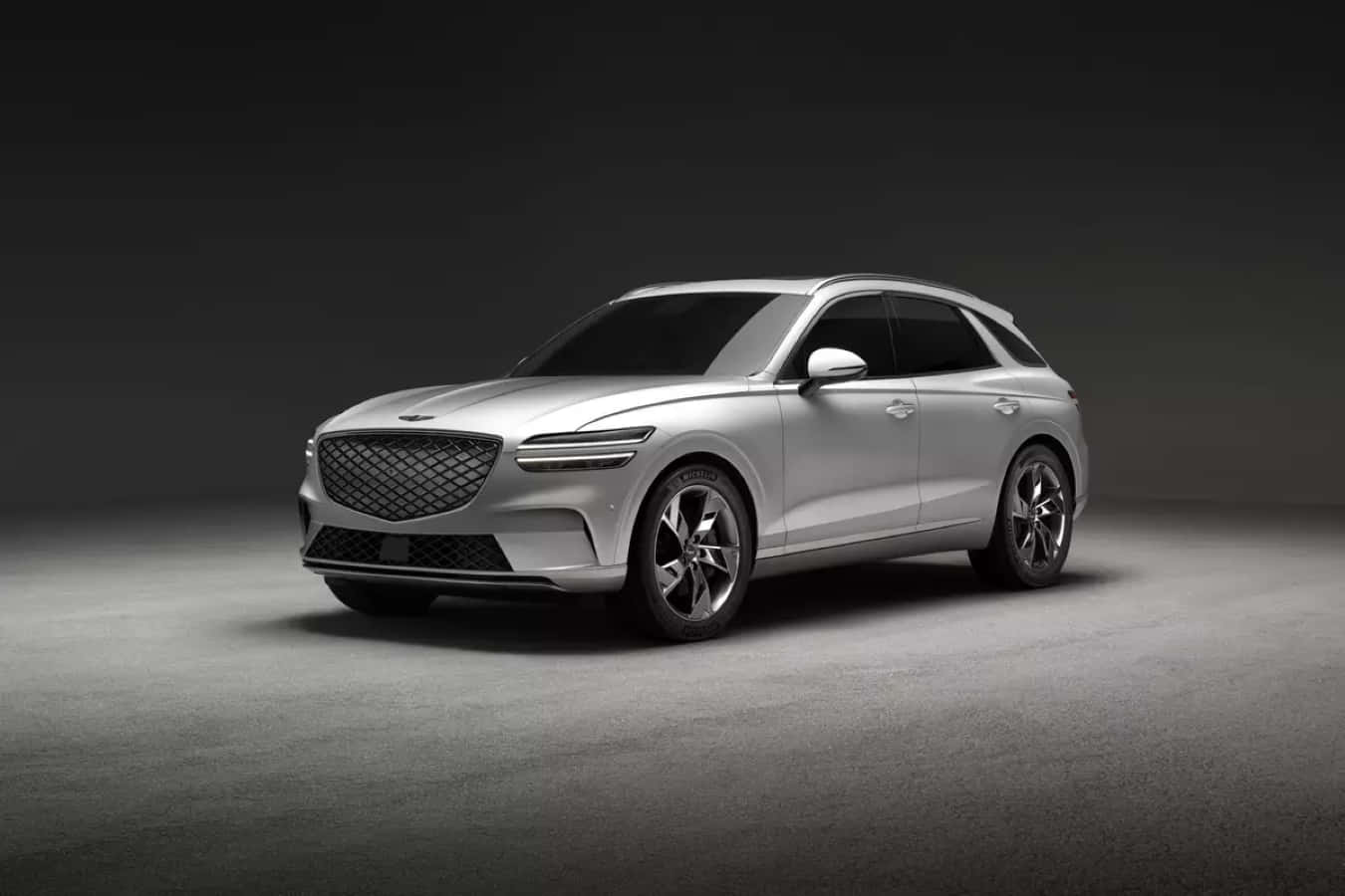 Genesis Gv70 桌布 壁紙 — 一輛時尚的銀色SUV，配備現代格柵和銳利線條，在戲劇性的工作室燈光下，以深色漸變背景展示。