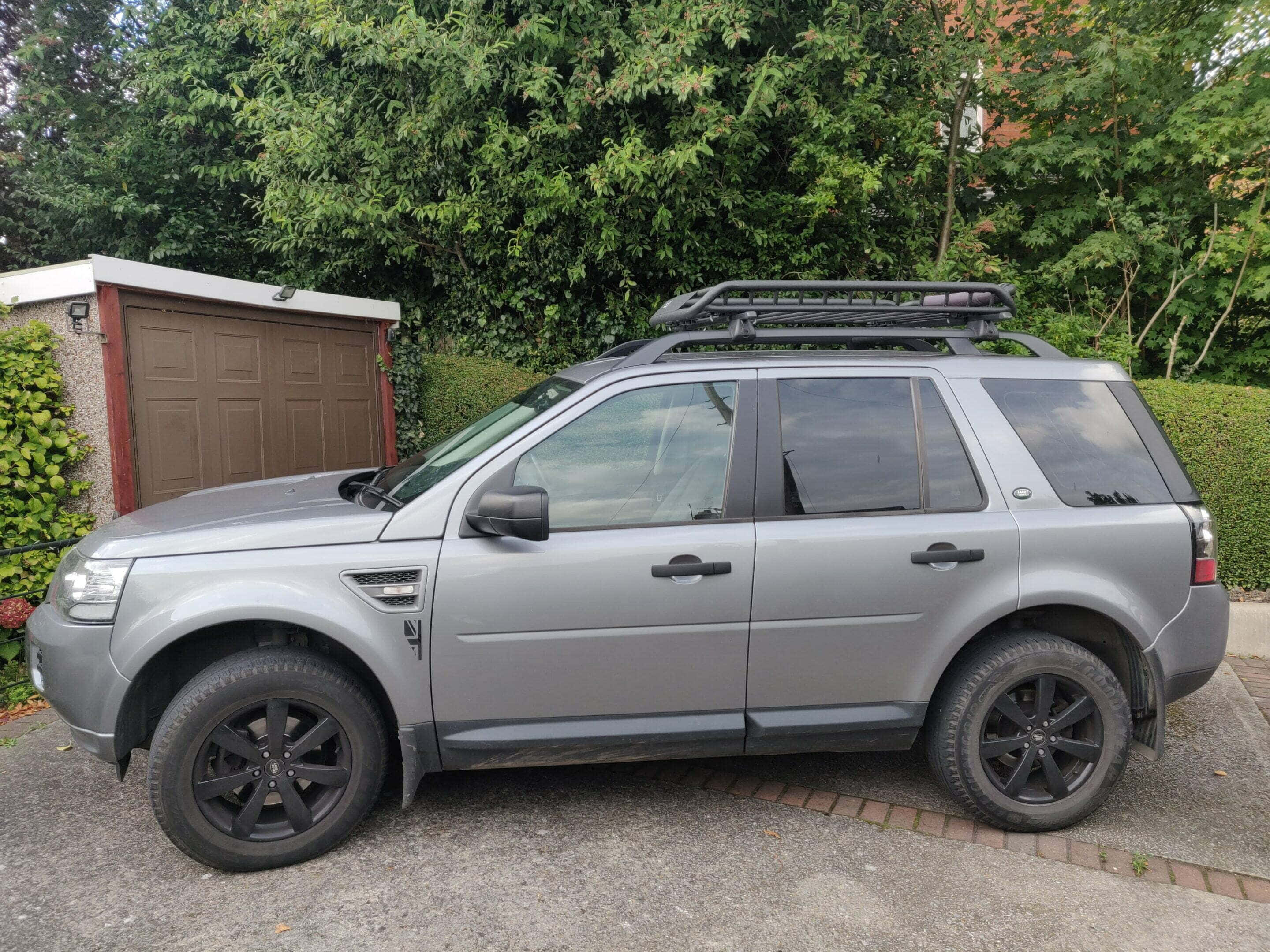 Land Rover Freelander 桌布 壁紙 — 一輛時尚的銀色SUV，配備黑色車輪和車頂行李架，停在鋪砌的車道上，旁邊是木車庫和茂密的綠色樹籬。