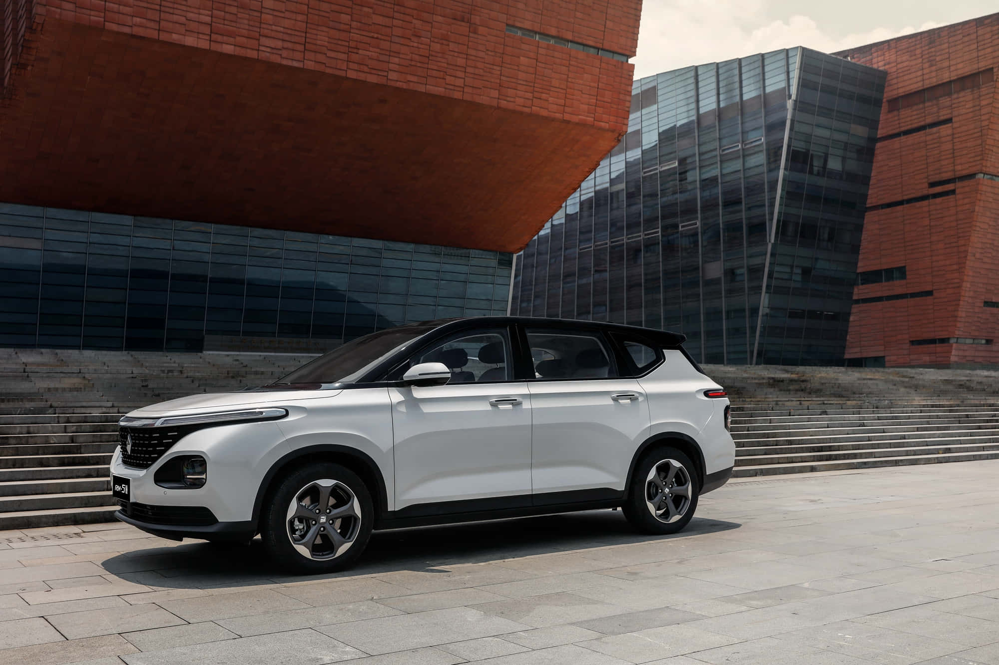 Baojun 桌布 壁紙 — 一輛時尚的銀色SUV停泊在石頭廣場上，前方是一座現代建築，擁有醒目的紅磚和玻璃外牆。