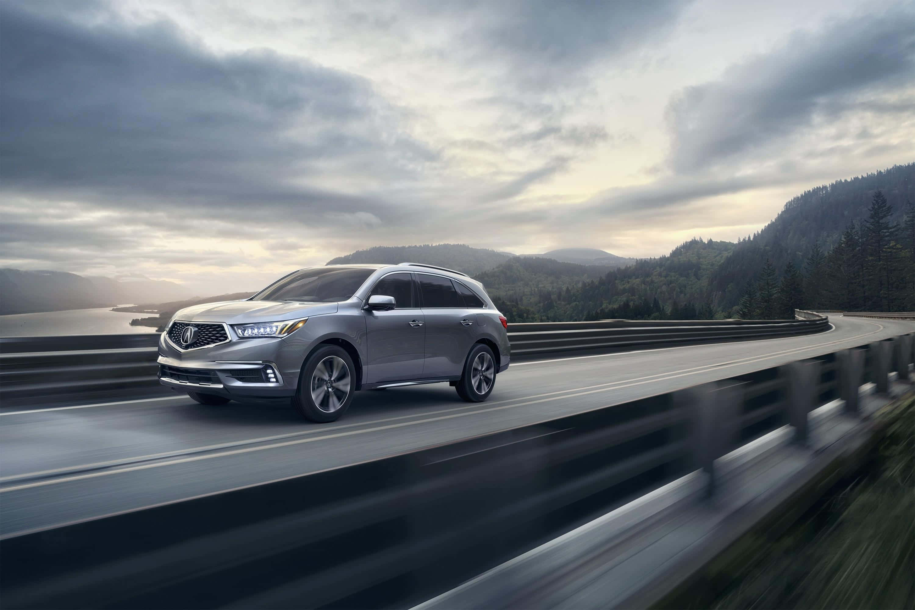 Acura Mdx 桌布 壁紙 — 一輛時尚的銀色SUV沿著蜿蜒的沿海公路行駛，背景是森林覆蓋的山脈和多雲的天空。