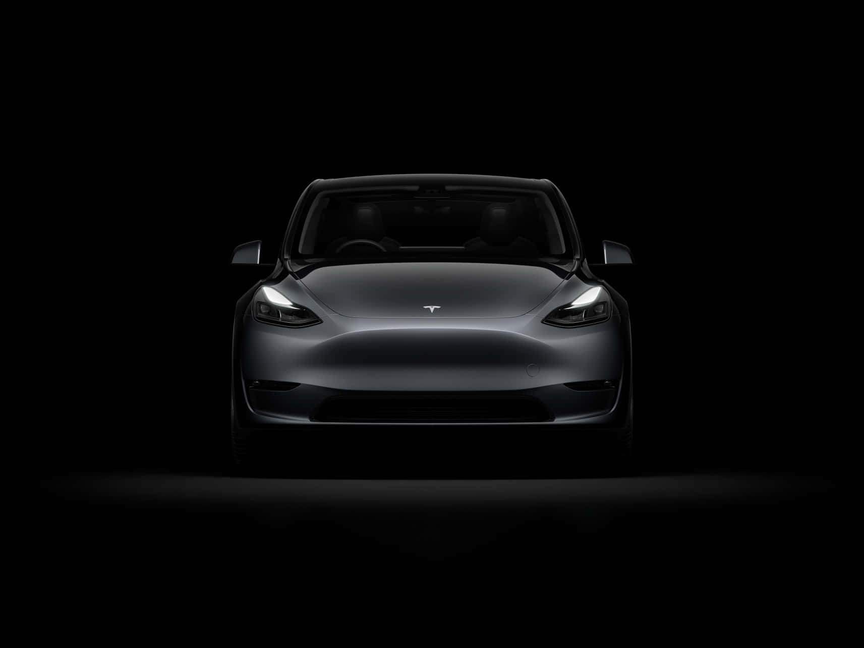 tesla 桌布 壁紙 — 一輛時尚的銀色Tesla Model 3正面朝前，車頭燈在漆黑的工作室背景中發光。