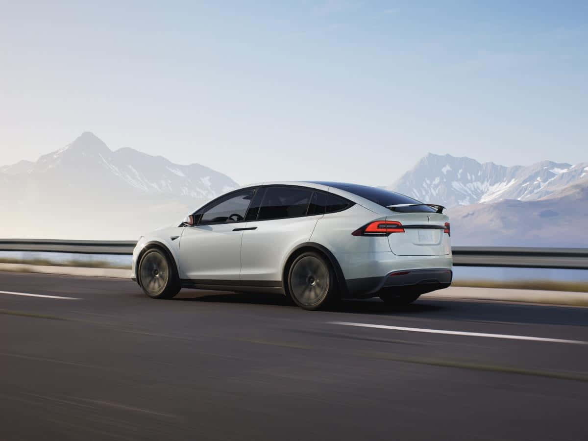 tesla 桌布 壁紙 — 一輛時尚的銀色Tesla Model X在高速公路上行駛，遠處朦朧的雪山在晴朗的天空下聳立。