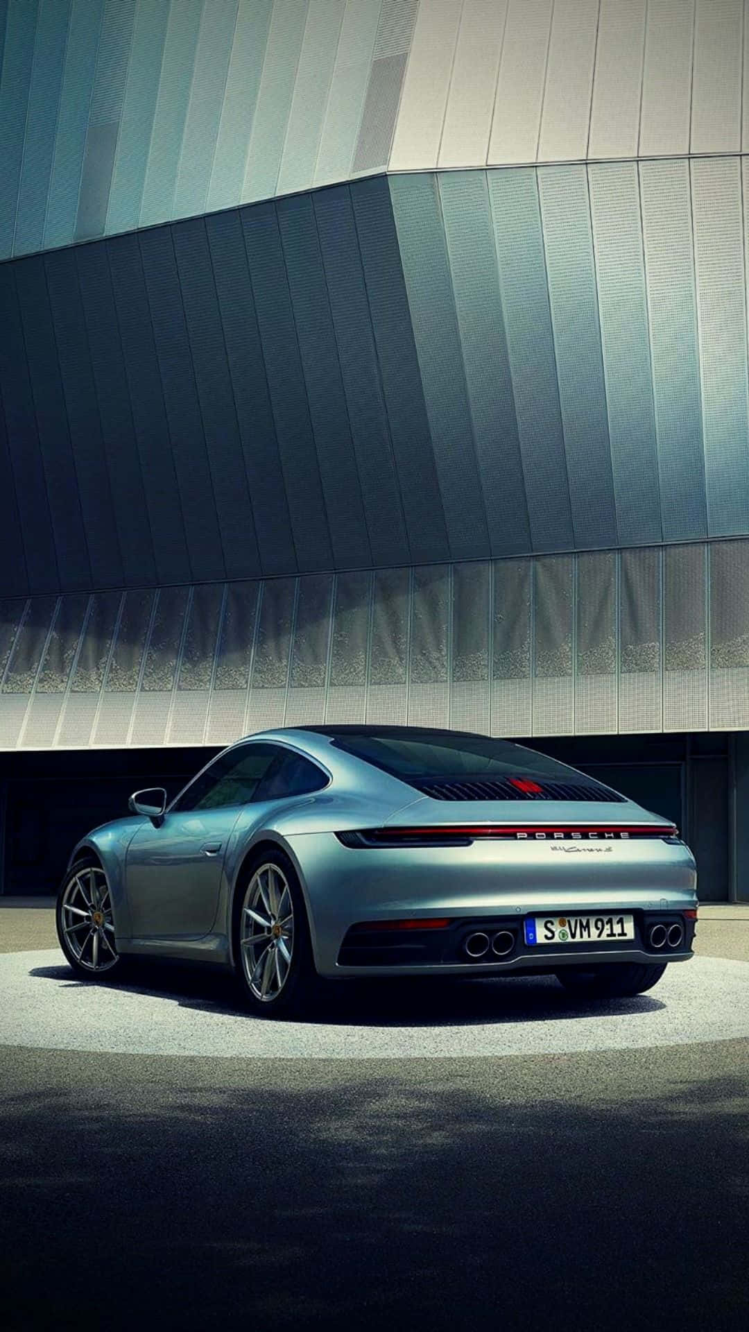 Porsche iPhone 桌布 壁紙 — 一輛時尚的銀色保時捷911跑車停在現代棱角分明的金屬外牆建築前。