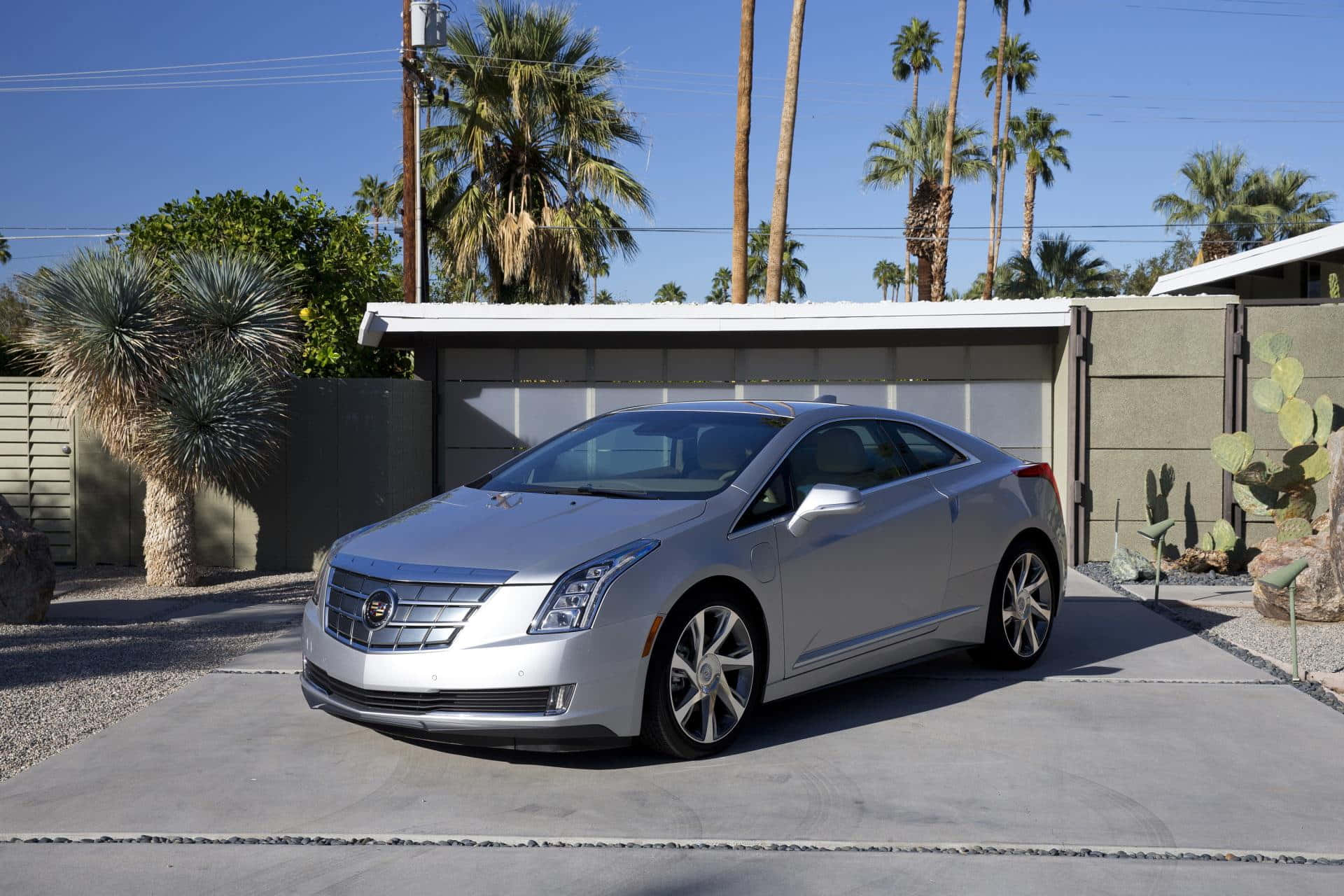 Cadillac Elr 桌布 壁紙 — 一輛時尚的銀色凱迪拉克ELR電動轎跑車停在現代房屋前，房屋周圍有棕櫚樹和仙人掌，天空湛藍。