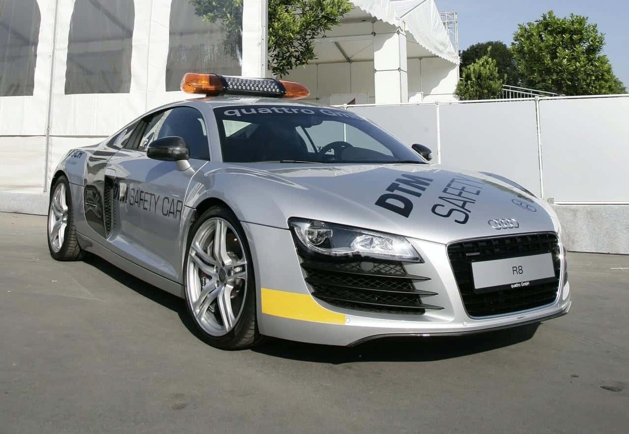 Safety Car 桌布 壁紙 — 一輛時尚的銀色奧迪R8安全車，帶有黃色點綴和車頂燈，停在白色帳篷和綠色樹木附近的混凝土場地上。