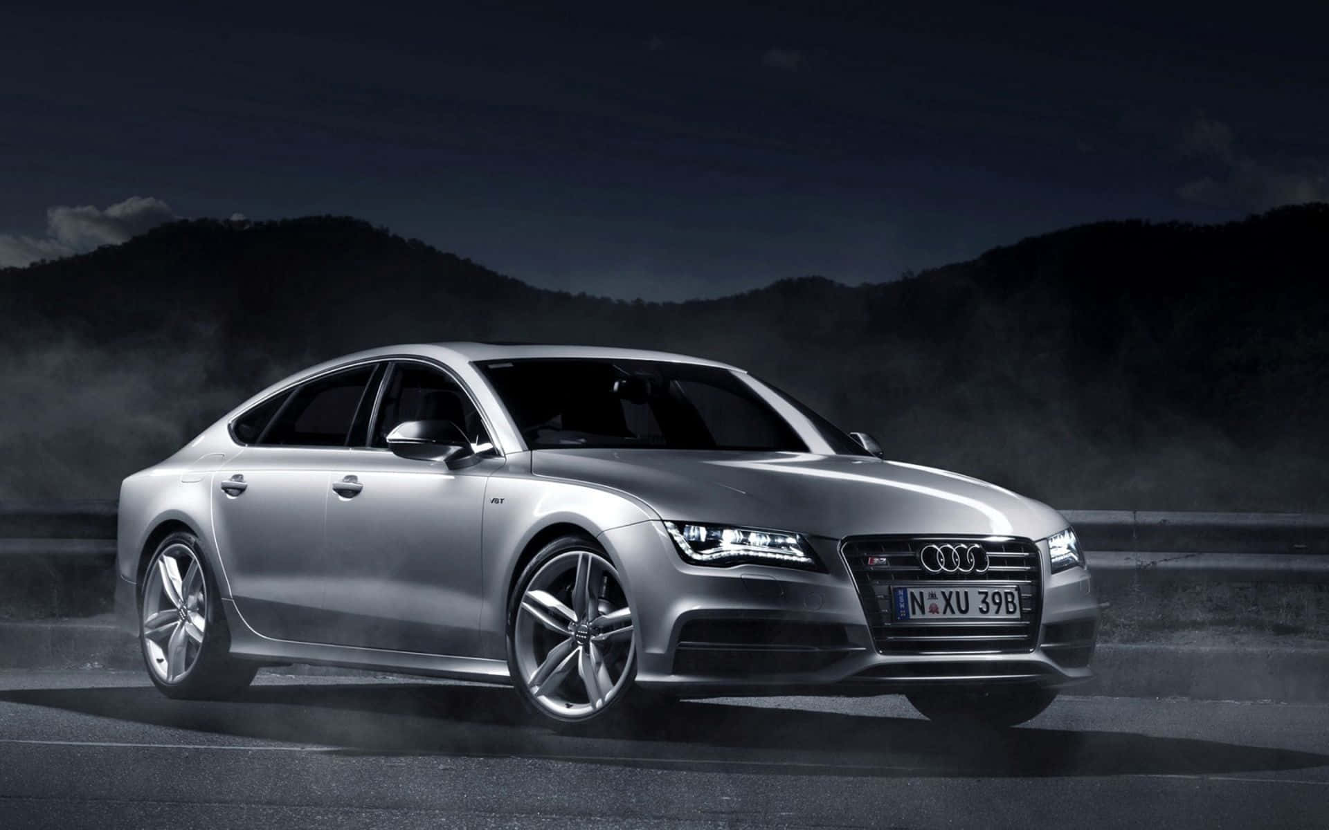 Audi S7 桌布 壁紙 — 一輛時尚的銀色奧迪轎車在霧夜中滑行，閃爍的車頭燈照亮了山巒背景。