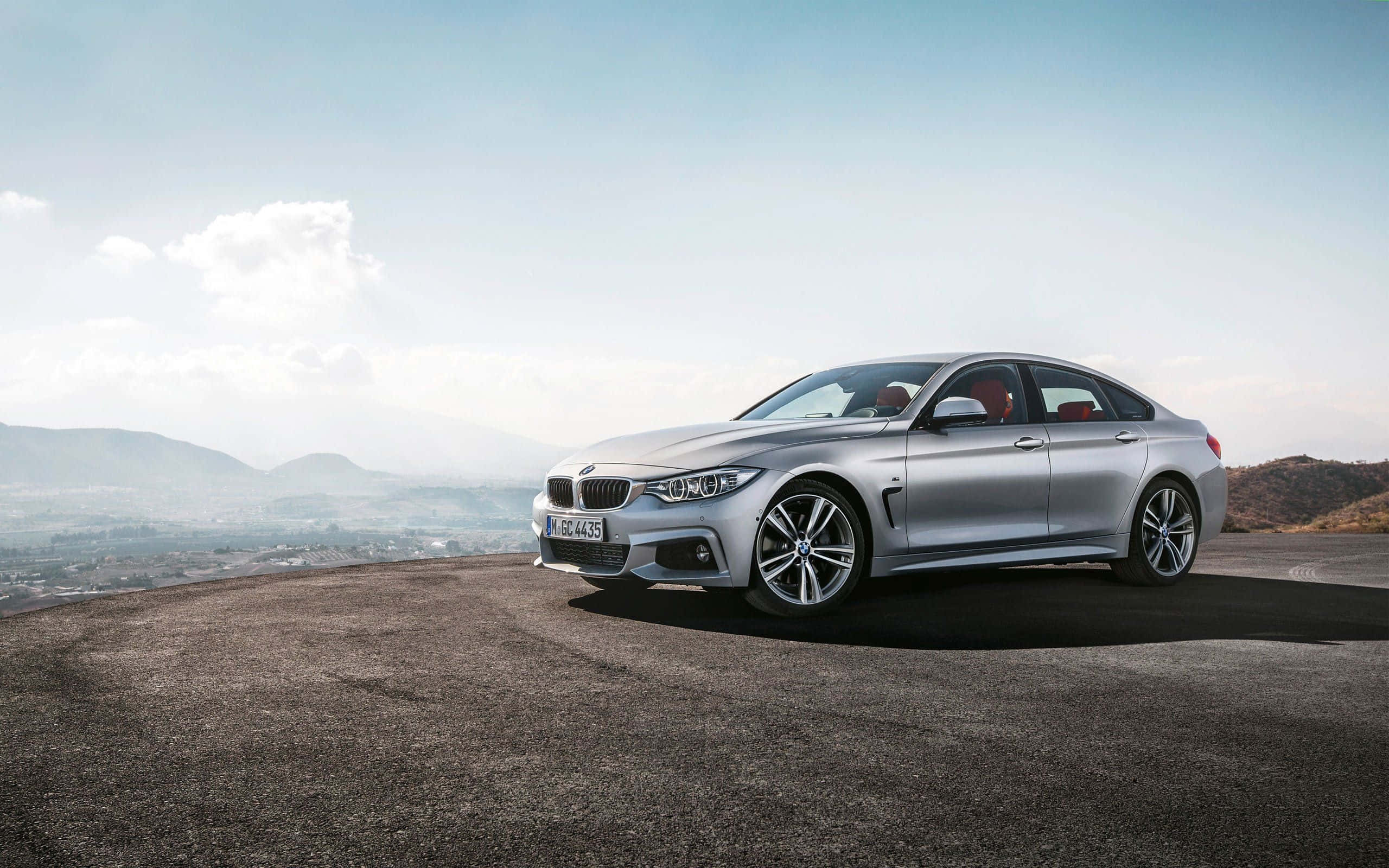 Bmw 4 Series 桌布 壁紙 — 一輛時尚的銀色寶馬房車停在高處觀景點，遠山和朦朧的城市天際線在明亮天空下。