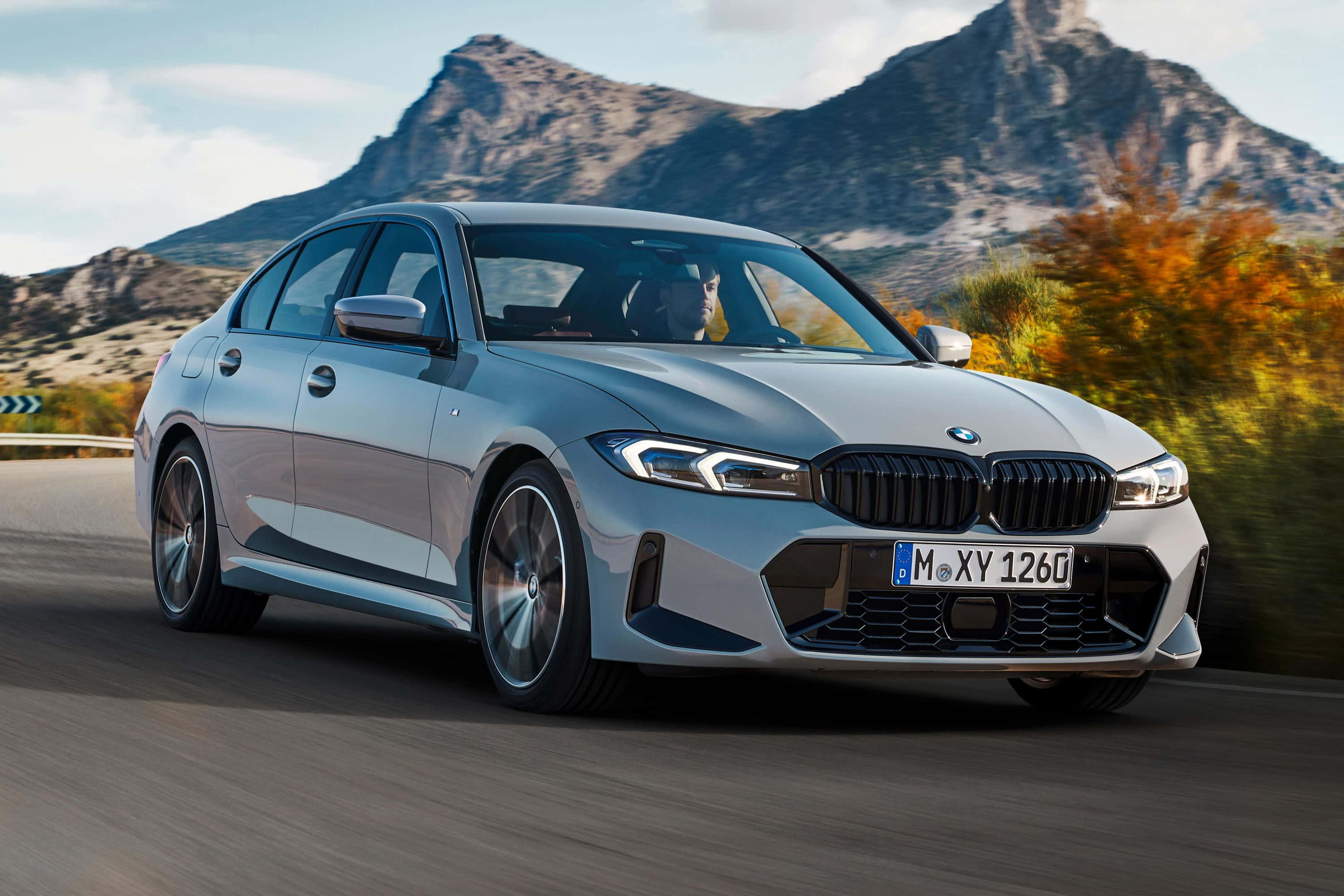 Bmw 3 Series 桌布 壁紙 — 一輛時尚的銀色寶馬房車在秋葉和崎嶇山峰背景下沿山路疾馳。