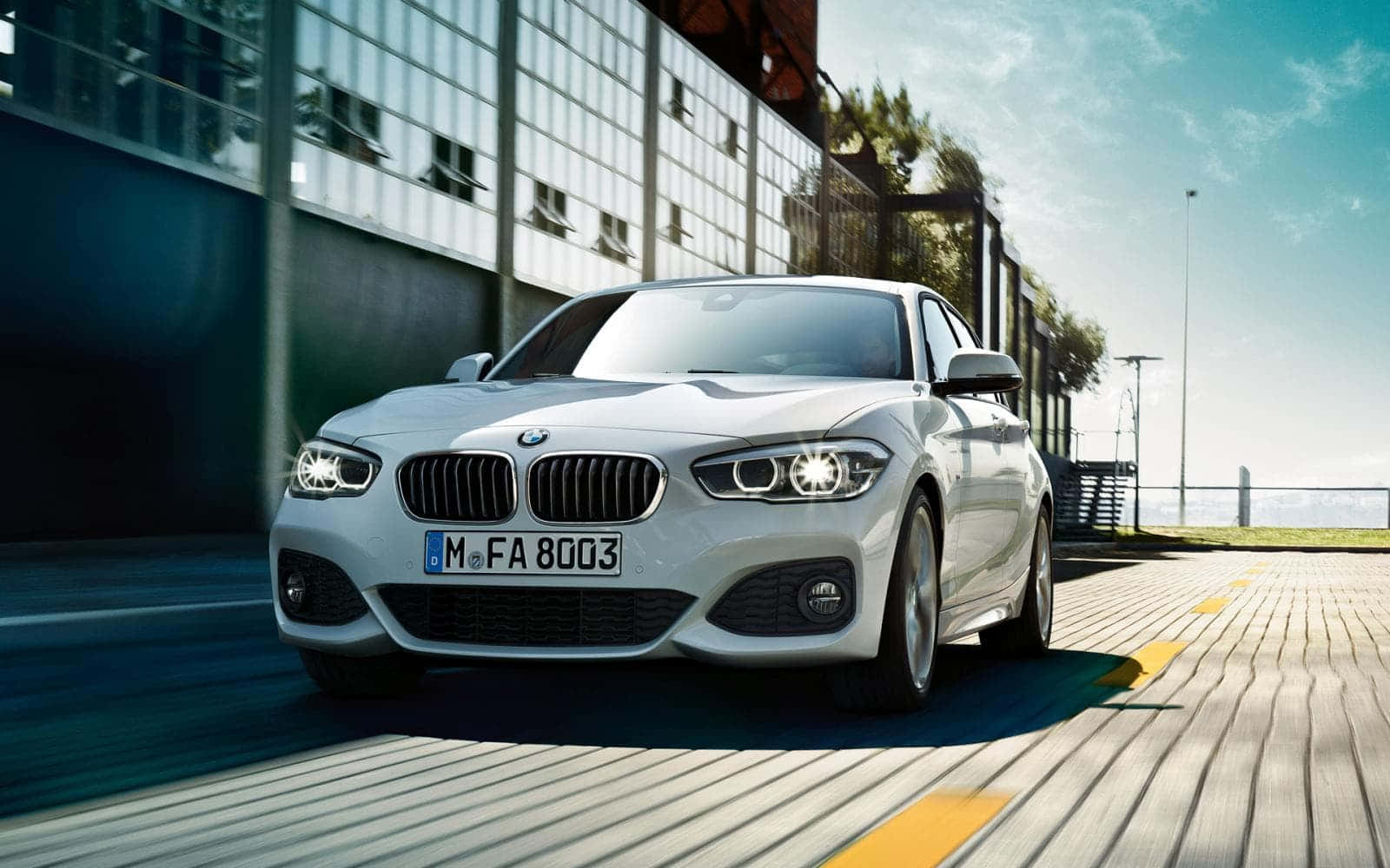 Bmw 1 Series 桌布 壁紙 — 一輛時尚的銀色寶馬房車在陽光普照的城市道路上飛馳，背景是現代化的玻璃建築。