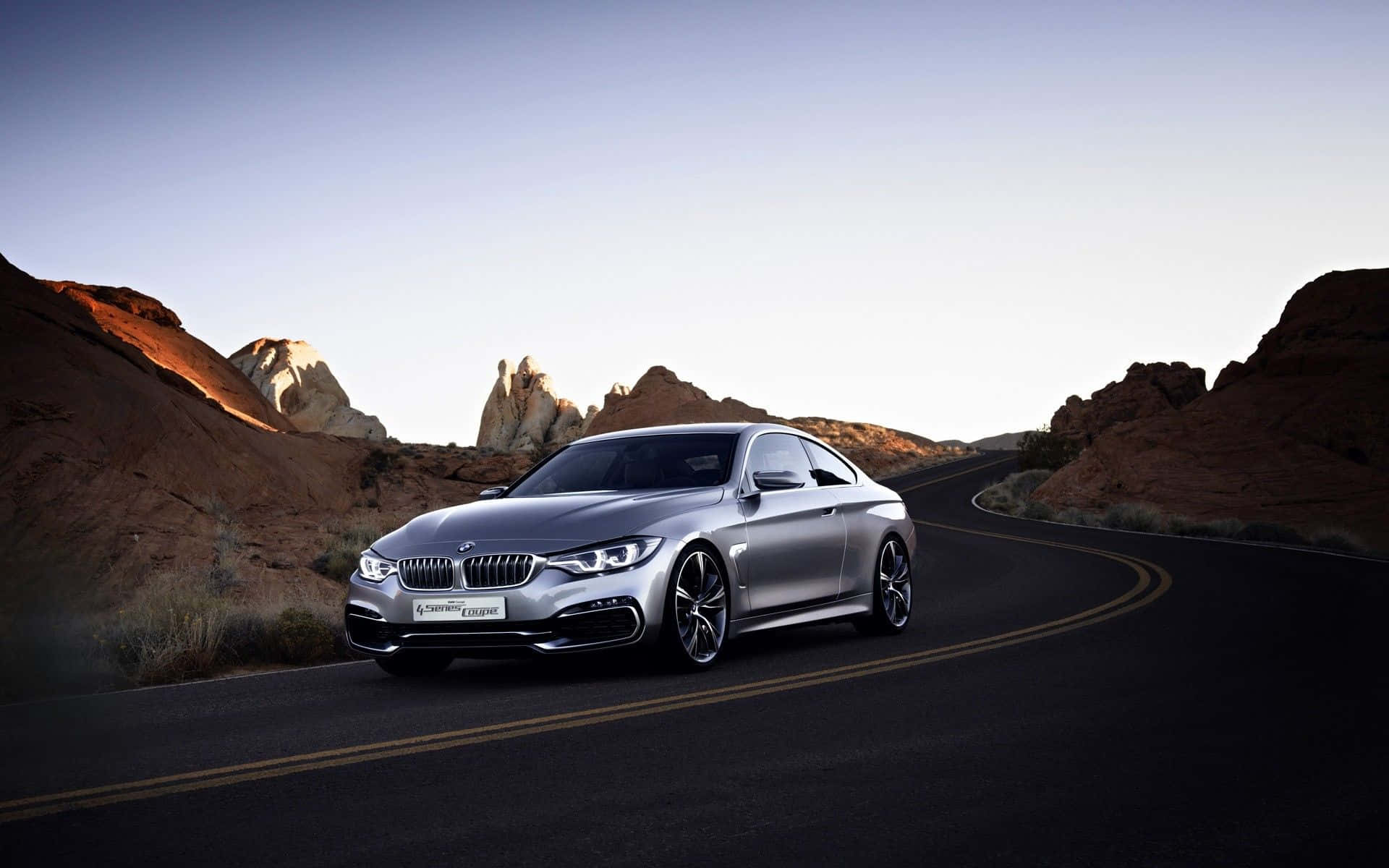 Bmw 4 Series 桌布 壁紙 — 一輛時尚的銀色寶馬轎跑車在柔和的暮色天空下，沿著兩旁紅色岩層的蜿蜒沙漠道路行駛。