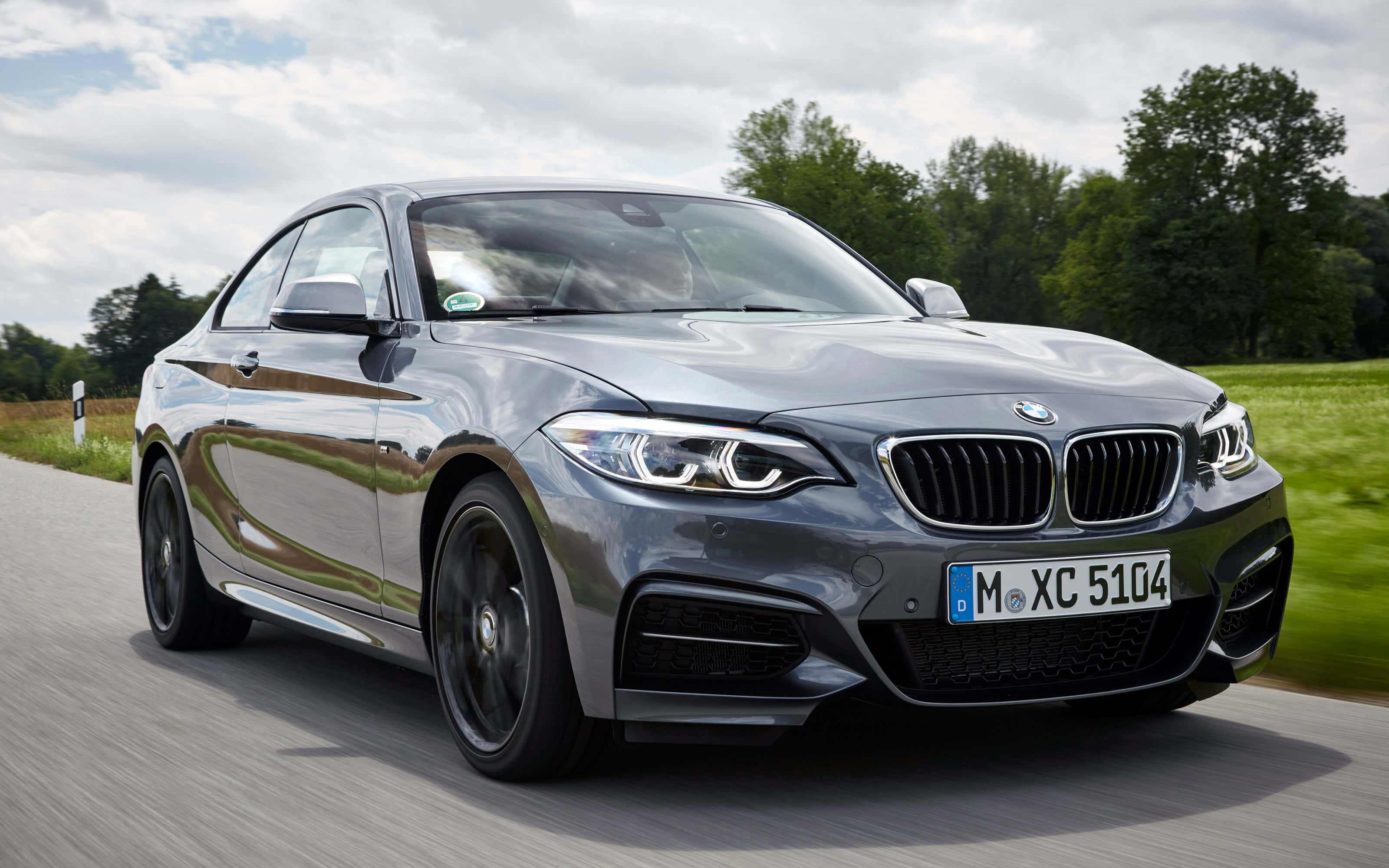 Bmw 2 Series 桌布 壁紙 — 一輛時尚的銀色寶馬轎跑車在陰天下的鄉村道路上疾馳，兩旁是綠色的田野和樹木。