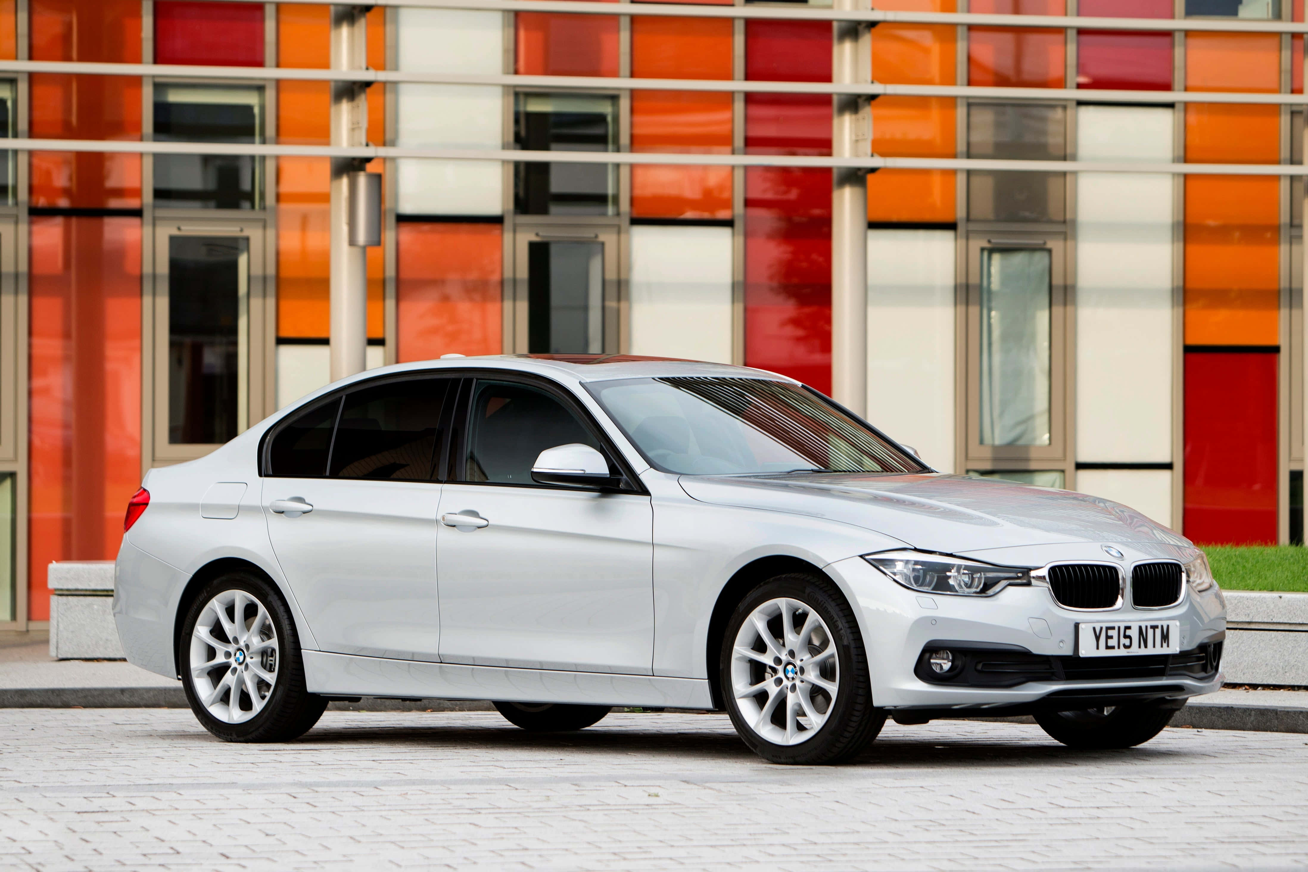 Bmw 3 Series 桌布 壁紙 — 一輛時尚的銀色寶馬轎車停泊在鋪砌的城市街道上，前面是一棟色彩繽紛的現代建築，帶有橙色、紅色和白色的面板。