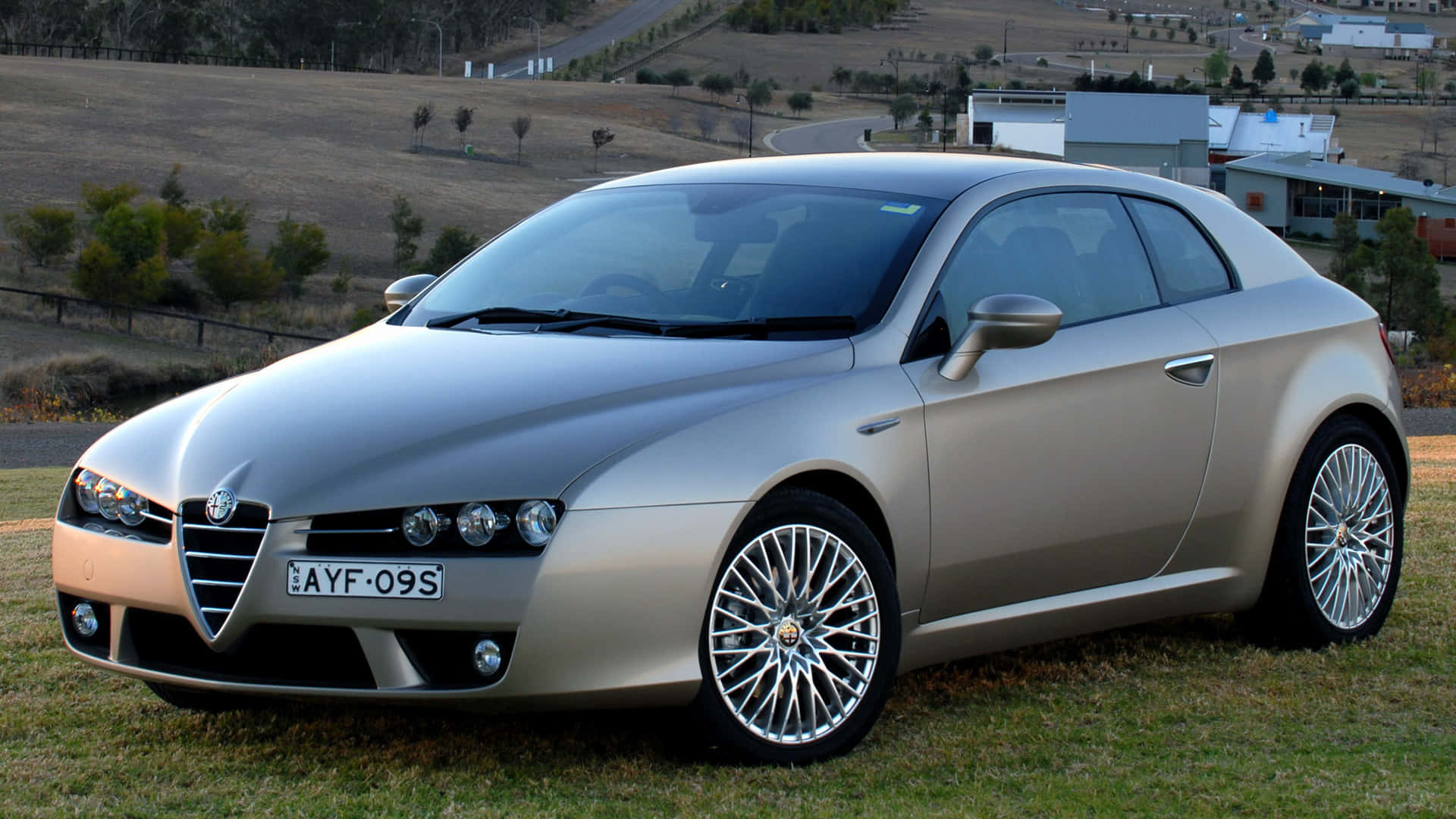 Alfa Romeo Brera 桌布 壁紙 — 一輛時尚的銀色愛快羅密歐Spider停在草地上，背景有起伏的山丘和遠處的建築物，在明亮的日光下。