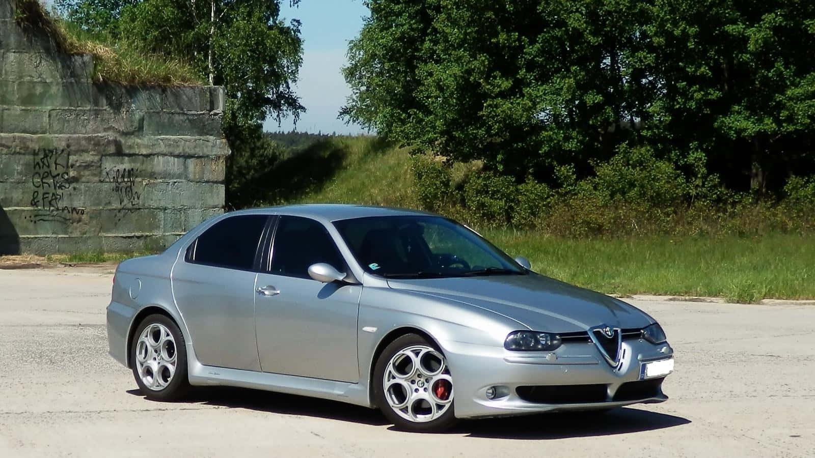 Alfa Romeo 156 桌布 壁紙 — 一輛時尚的銀色愛快羅密歐房車停泊在混凝土停車場，旁邊是長滿青苔的石牆和翠綠樹木，在明亮的日光下。