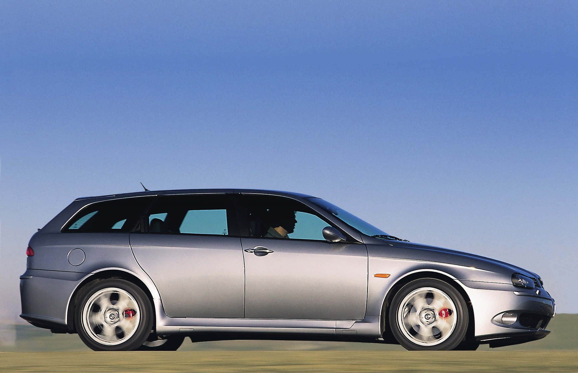 Alfa Romeo 156 桌布 壁紙 — 一輛時尚的銀色旅行車在晴朗的藍天下駛過草地，車內可見司機。