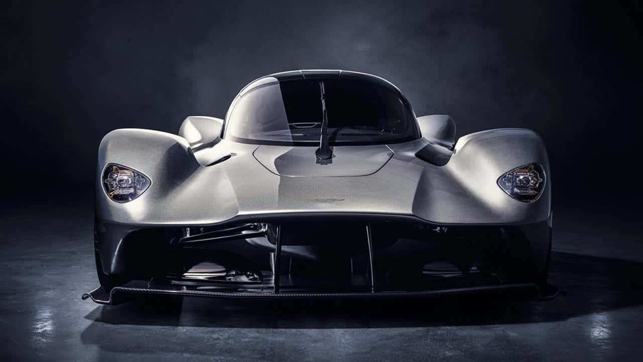 Aston Martin Valkyrie 桌布 壁紙 — 一輛時尚的銀色未來主義跑車，配備發光車頭燈，在昏暗的攝影棚中，在戲劇性的燈光下直視前方。