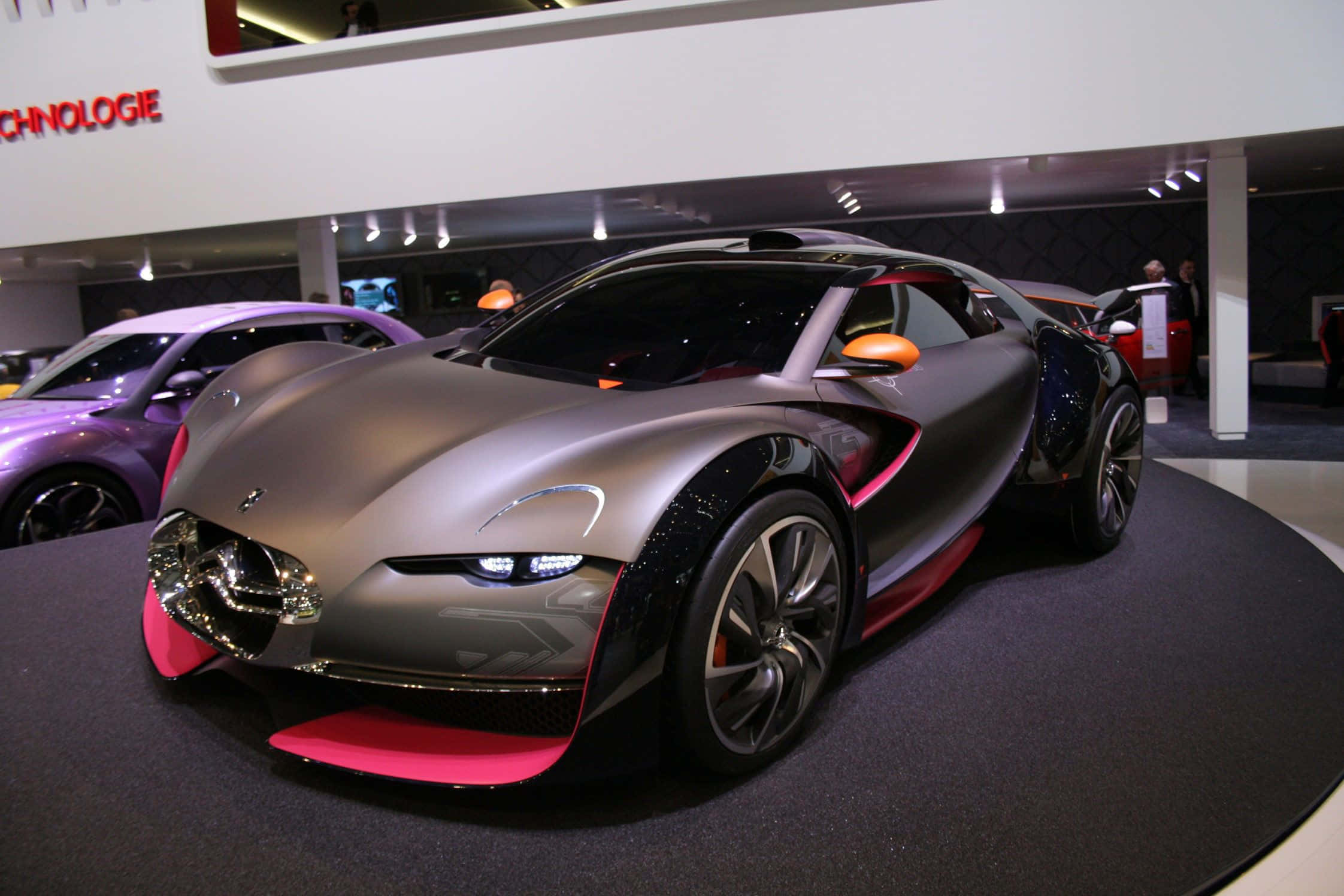 Citroen Ds Survolt 桌布 壁紙 — 一輛時尚的銀色未來感跑車帶有粉紅色點綴，在明亮的燈光下於黑色平台上展示於車展中。