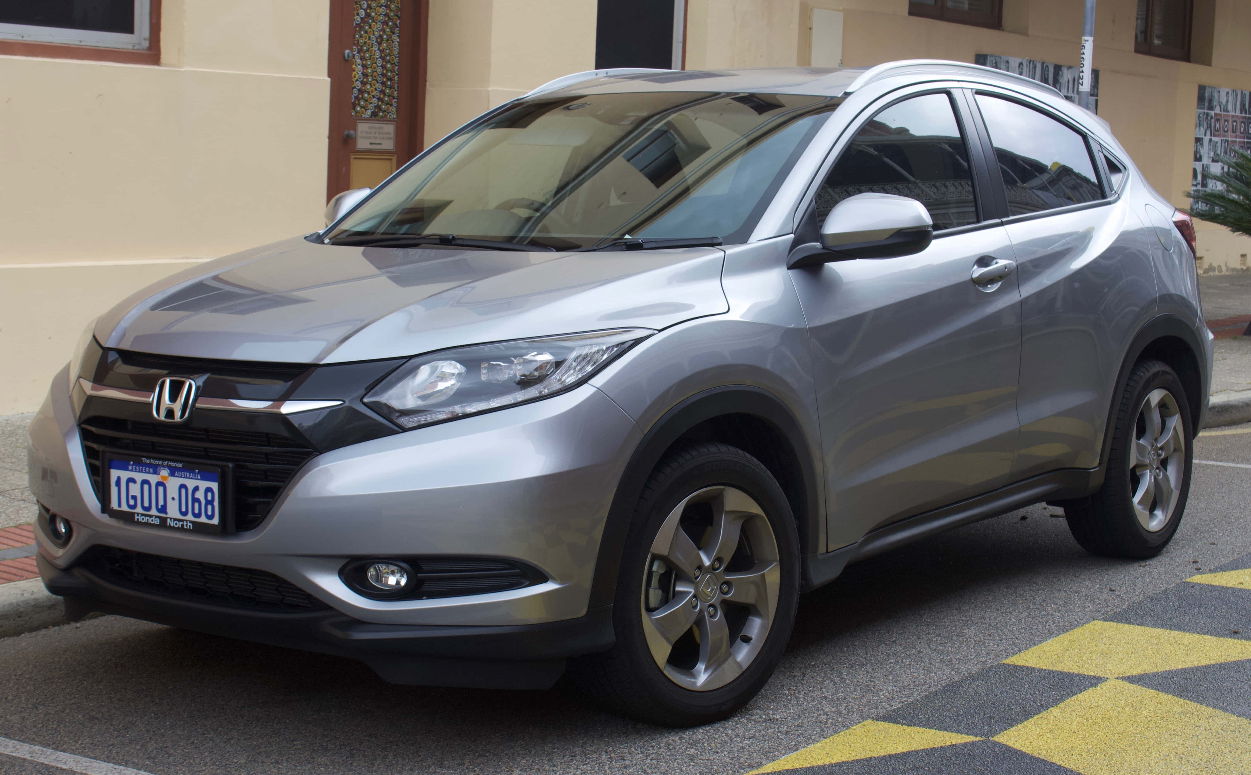 Honda Hr-v 桌布 壁紙 — 一輛時尚的銀色本田HR-V停在一條有黃色棋盤格路面的街道上，旁邊是一棟米色建築。
