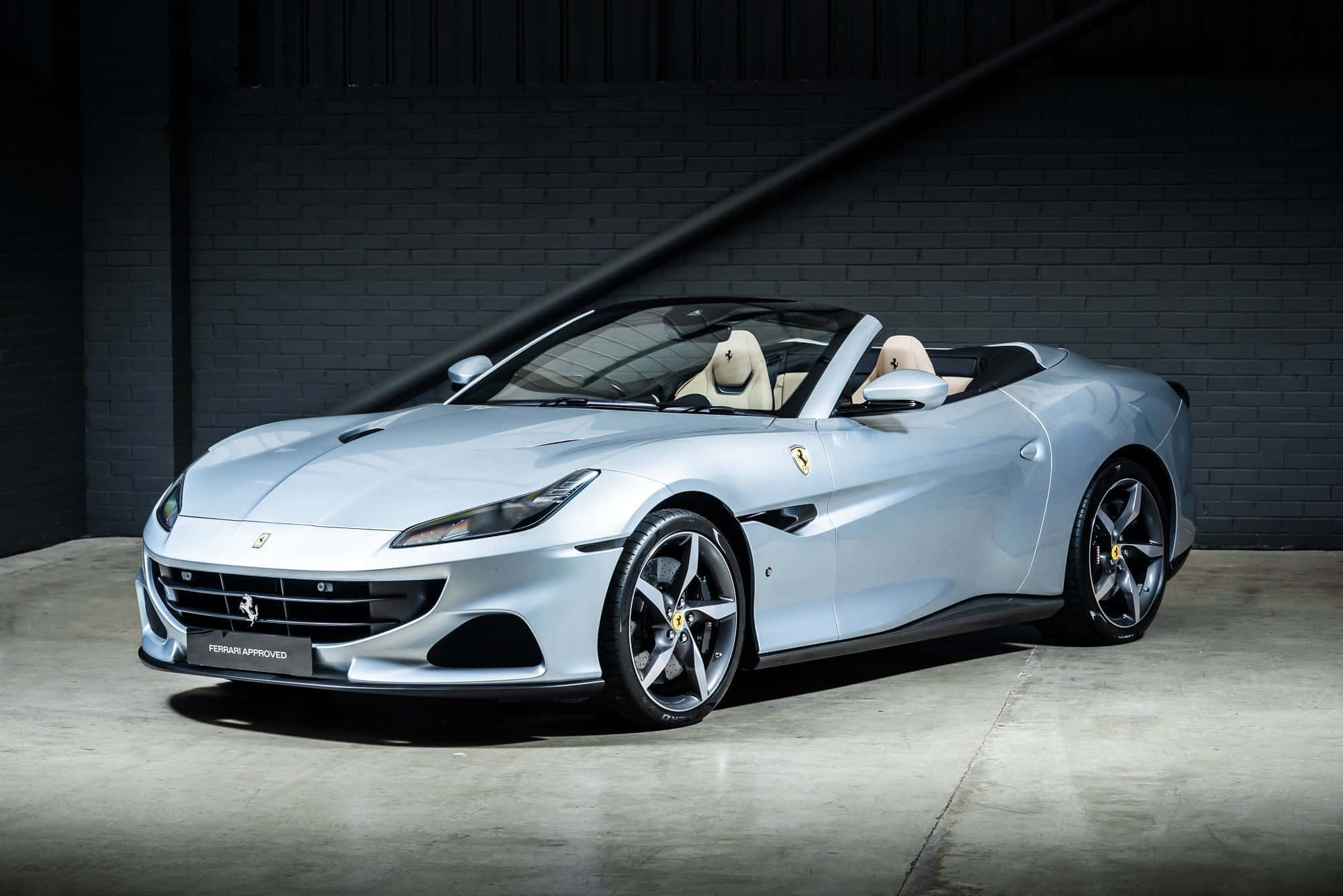 Ferrari Portofino 桌布 壁紙 — 一輛時尚的銀色法拉利Portofino開篷車，配備米色皮革座椅，停放在光線昏暗的工業車庫中，車庫有裸露的磚牆。
