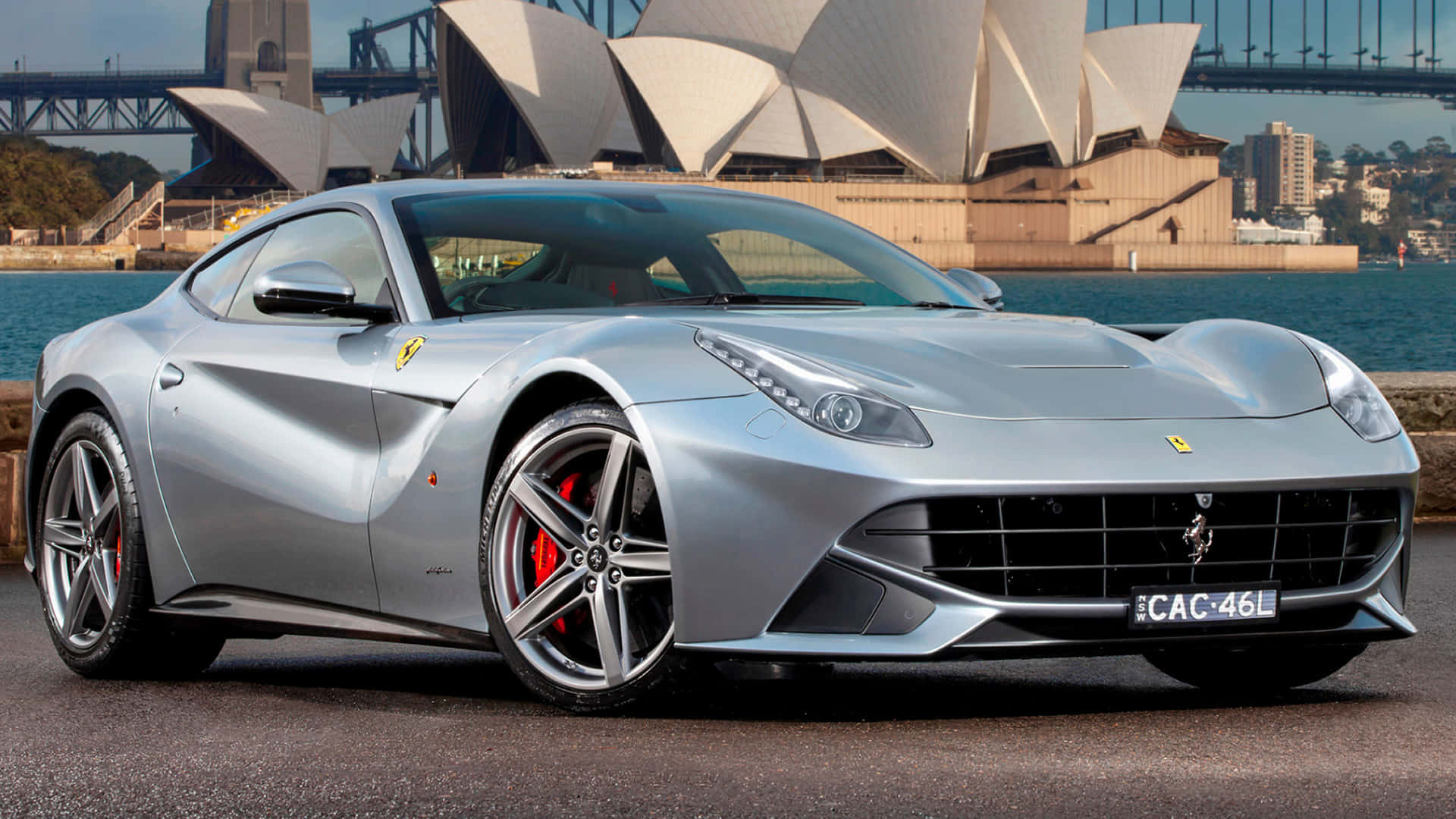 Ferrari F12 Berlinetta 桌布 壁紙 — 一輛時尚的銀色法拉利跑車停在水邊，背景是標誌性的悉尼歌劇院和海港大橋。