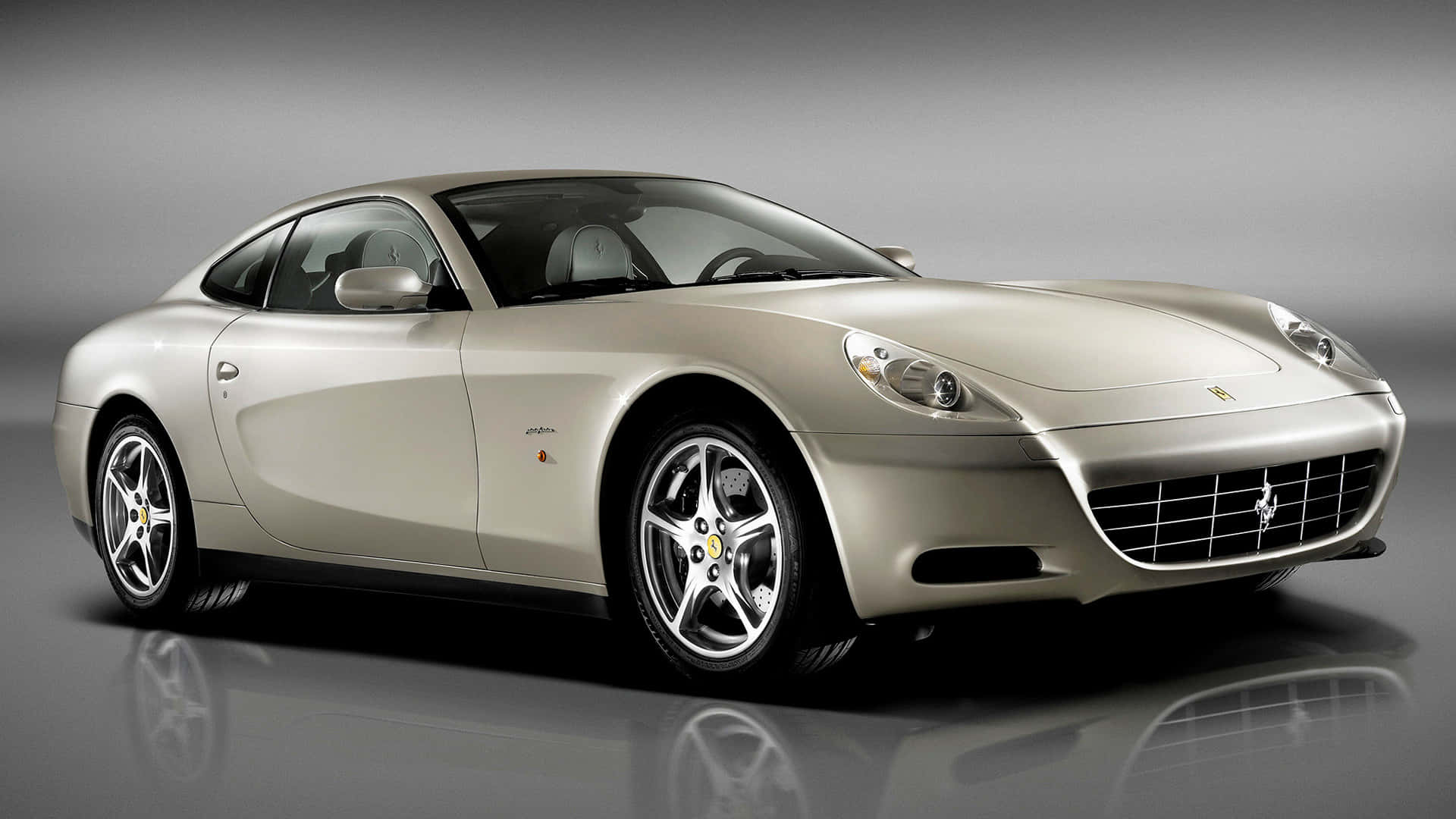Ferrari 612 Scaglietti 桌布 壁紙 — 一輛時尚的銀色法拉利轎跑車在攝影棚燈光下閃閃發光，搭配反射地板和漸變灰色背景。