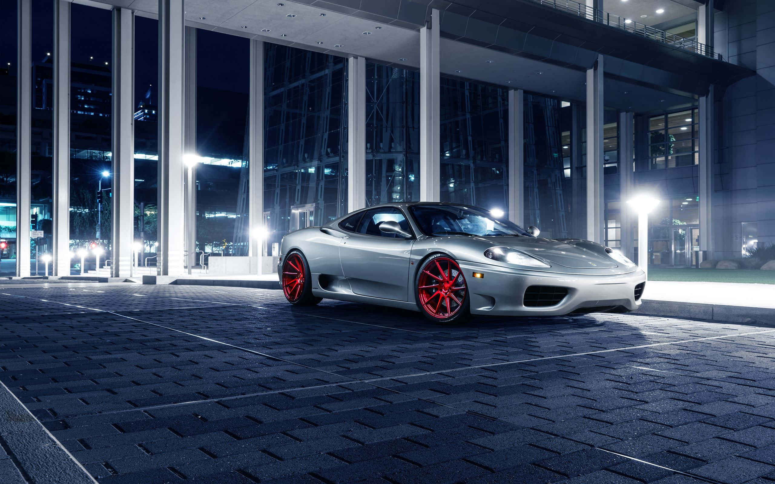 Ferrari 360 Modena 桌布 壁紙 — 一輛時尚的銀色法拉利配以鮮紅色輪圈，夜晚停泊在現代玻璃建築前。