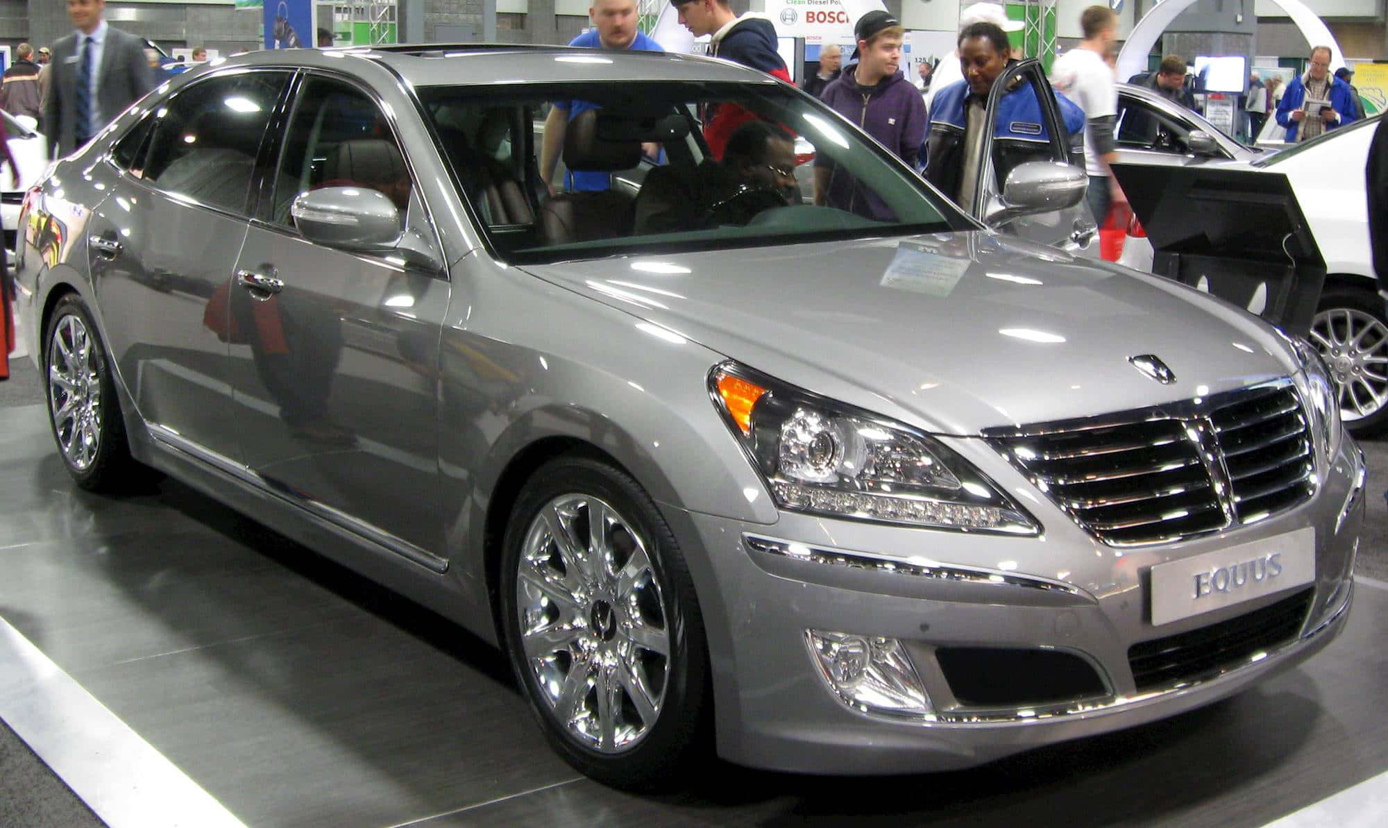 Hyundai Equus 桌布 壁紙 — 一輛時尚的銀色現代Equus轎車在明亮的展覽燈光下閃閃發光，與會者在車展上欣賞它。